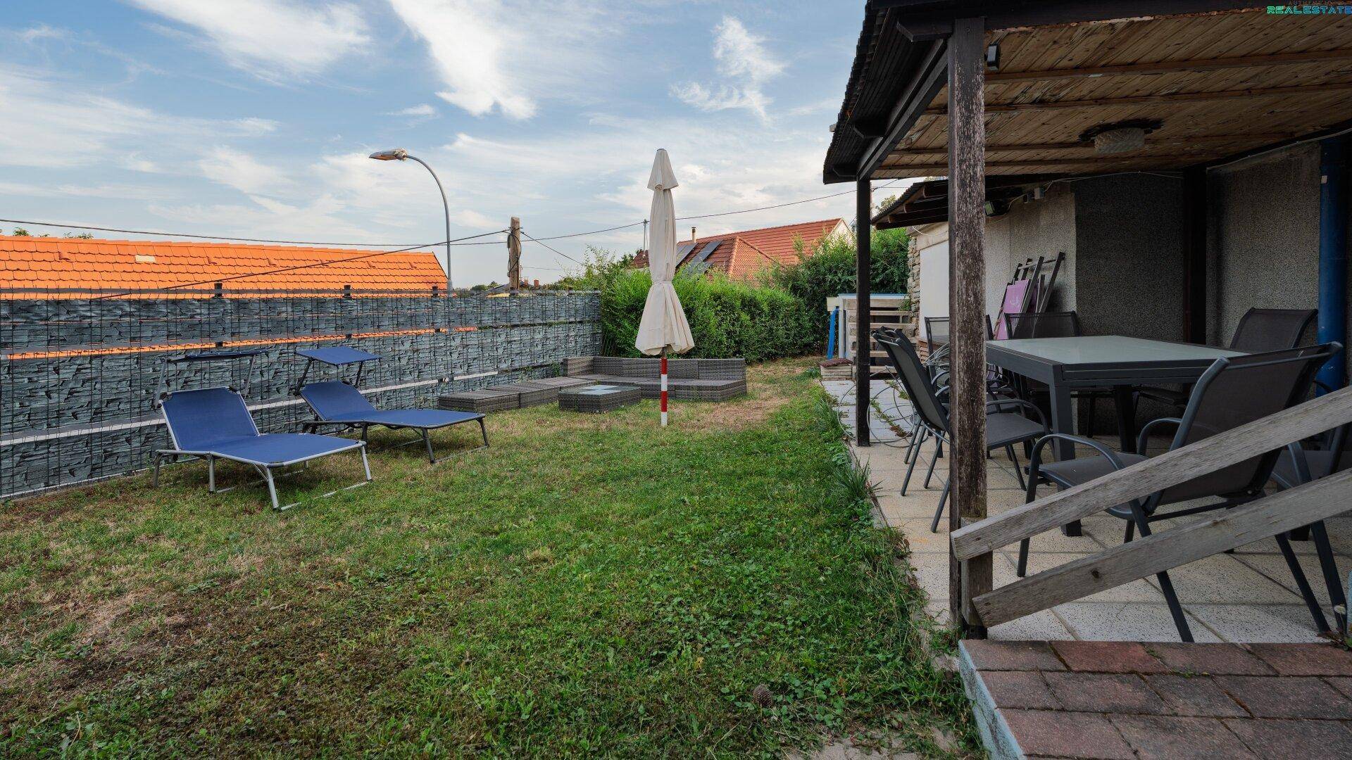 Gartenbereich mit überdachter Terrasse, Rasenfläche und Liegestühlen.