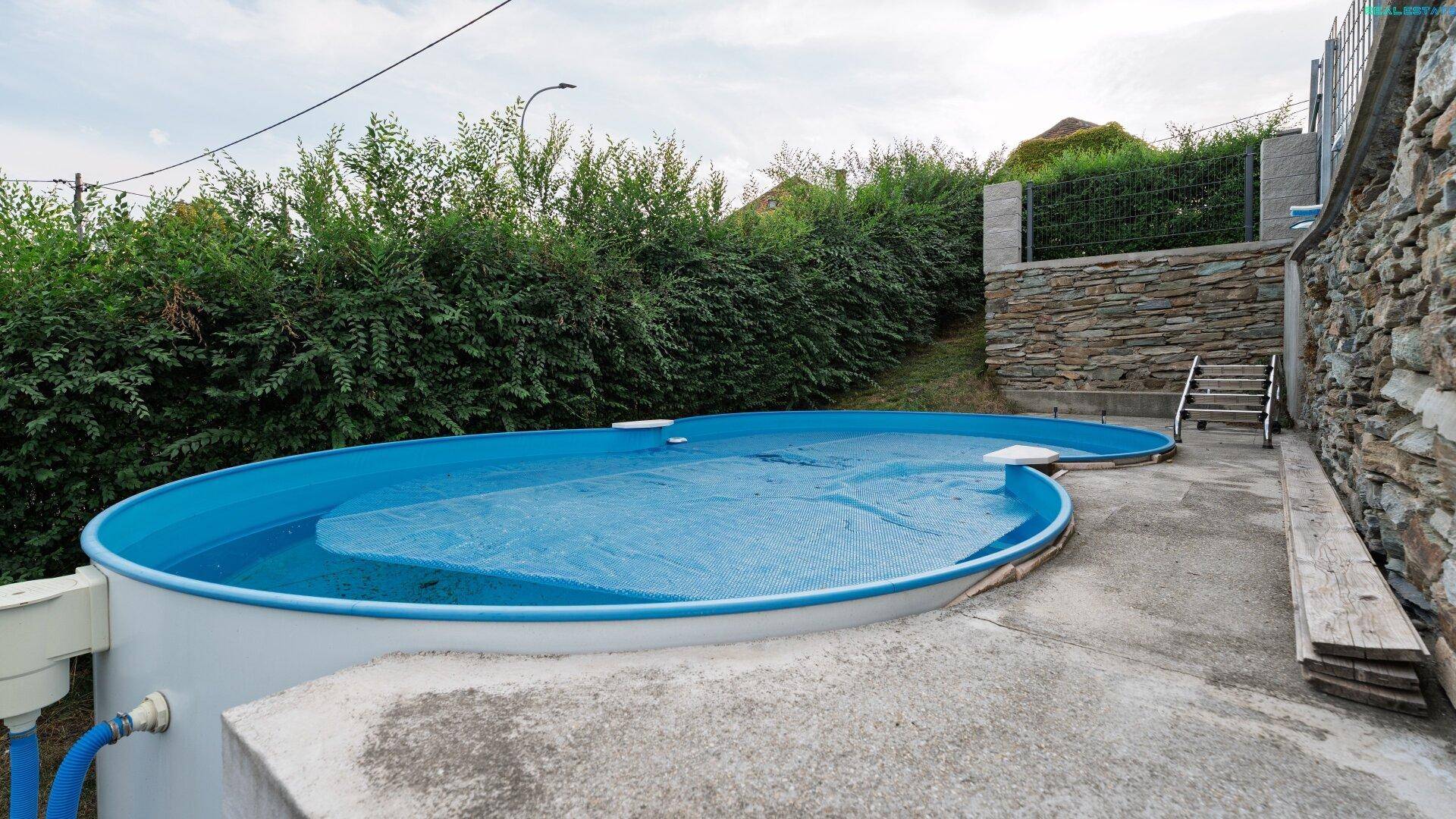 Ovaler Aufstellpool im Gartenbereich, umgeben von Beton und einer Steinmauer.