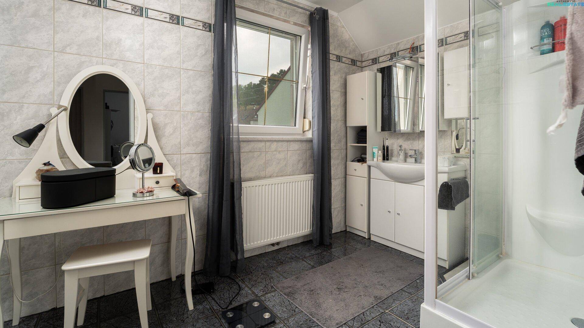 Gefliestes Badezimmer mit Duschkabine, Waschtisch und Schminktisch am Fenster.
