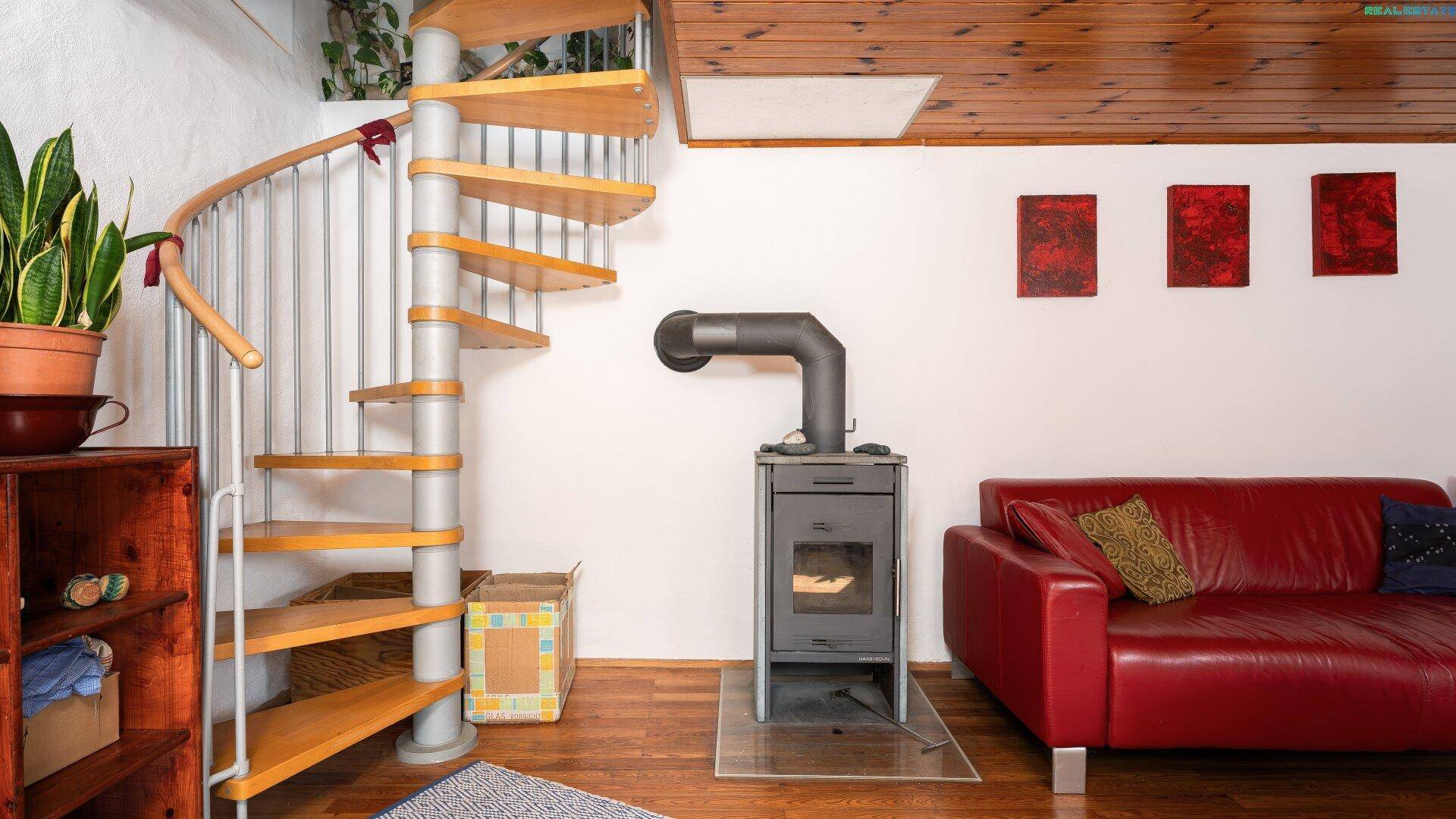 Detailansicht der Holzwendeltreppe und des Holzofens im Wohnbereich mit rotem Sofa.