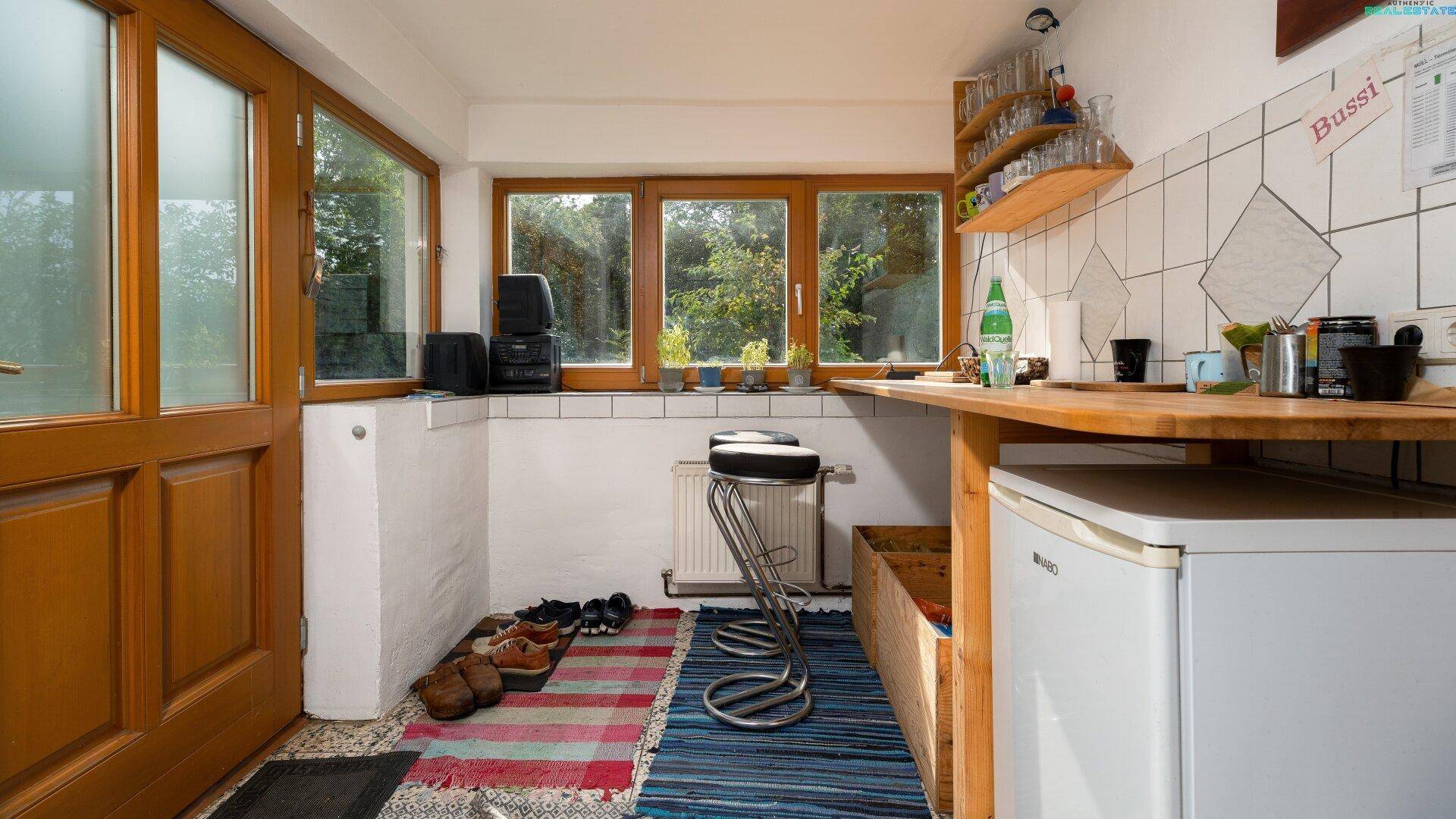 Helle Küche mit Holztür, Fenstern, kleinem Kühlschrank und gefliesten Wänden.