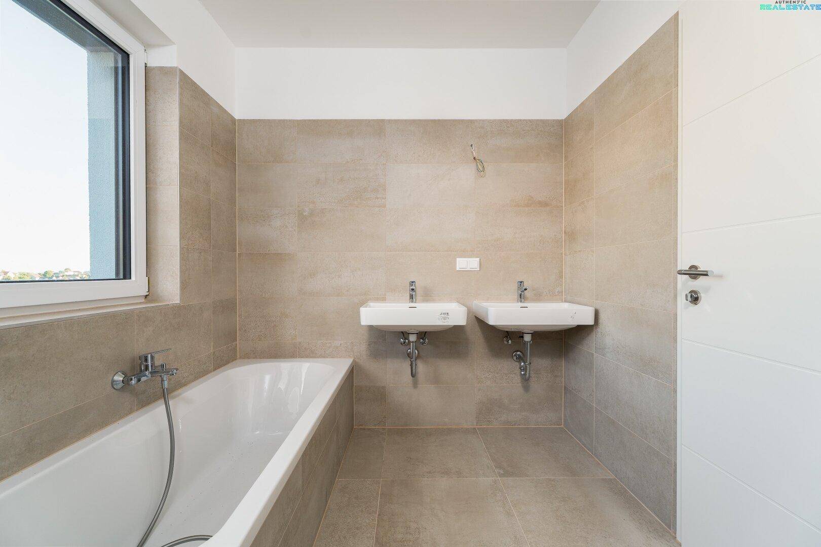 Helles Badezimmer mit Badewanne, zwei Waschbecken und großem Fenster für Tageslicht.