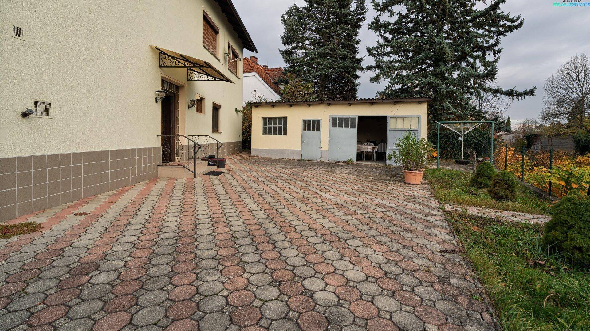 Gepflasterte Auffahrt zum Haus und zur Garage, umgeben von Grünflächen und Bäumen.