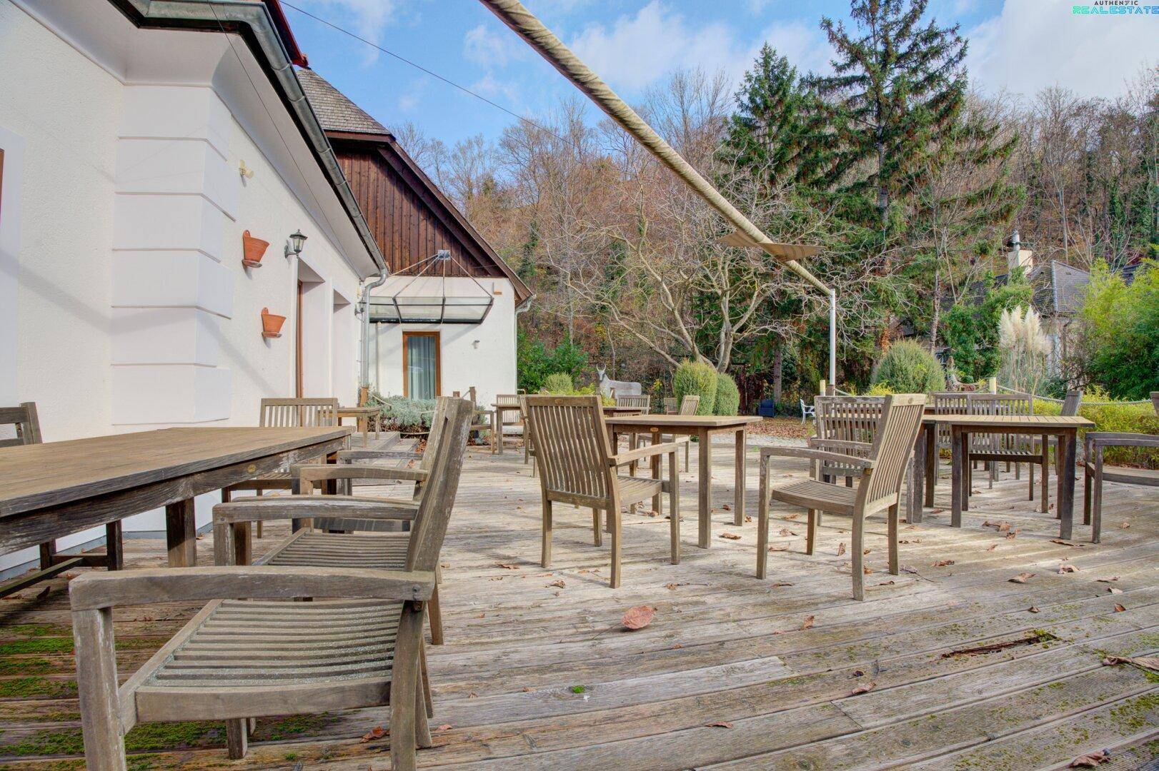 Weitläufige Holzterrasse mit vielen Tischen und Stühlen, umgeben von Bäumen und Sträuchern.