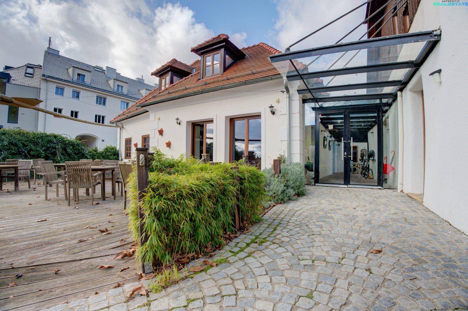 Eingangsbereich des Gebäudes mit Glasüberdachung und angrenzender Holzterrasse, umgeben von Grün.