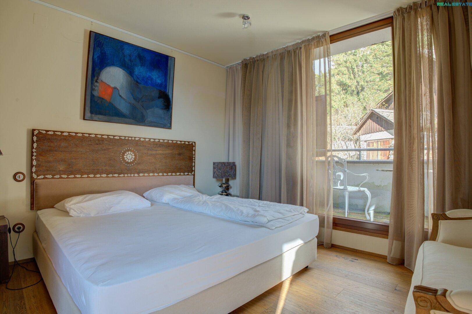Helles Schlafzimmer mit Doppelbett, Holzboden und Zugang zu einem kleinen Balkon mit Blick ins Grüne.