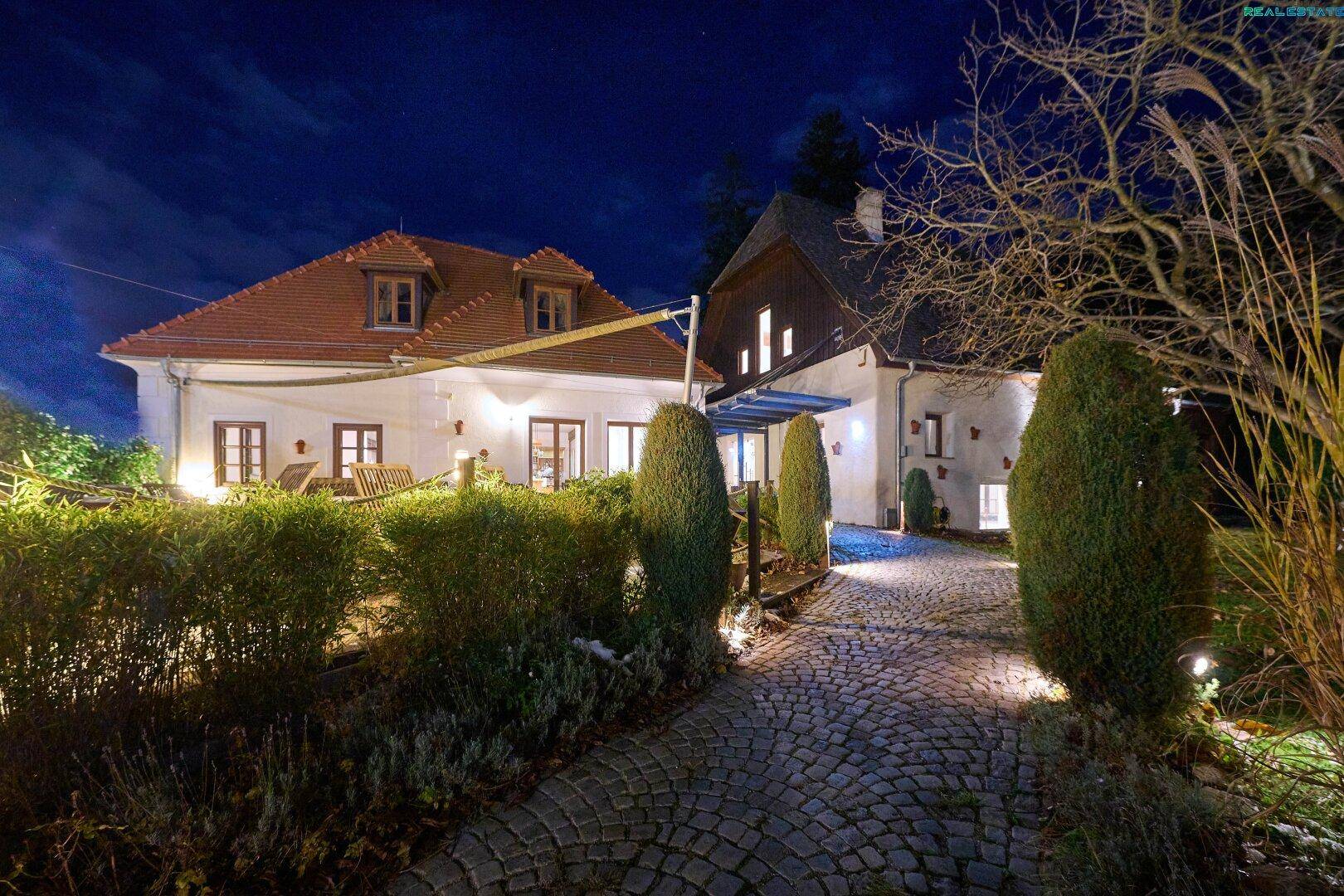 Beleuchtete Gebäudeansicht bei Nacht mit gepflasterter Auffahrt und gepflegter Gartenanlage.