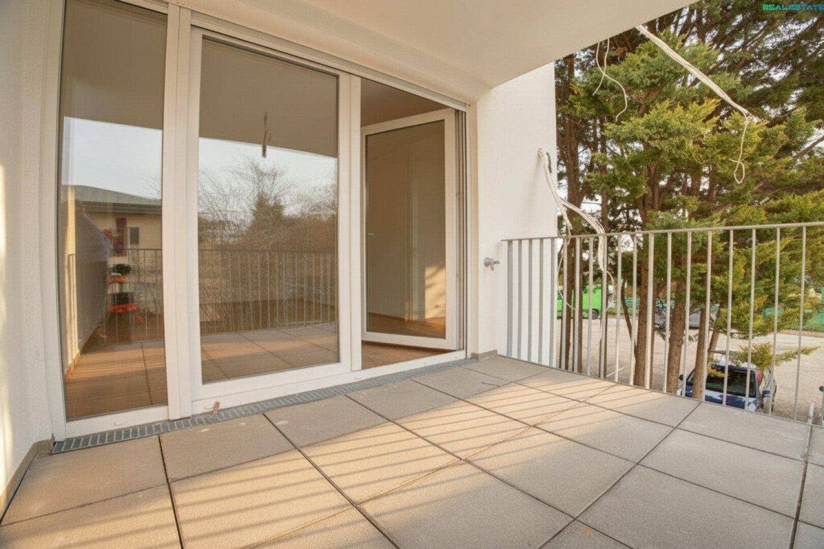 Heller Balkon mit Fliesenboden und Zugang durch große Schiebetüren, umgeben von Bäumen.