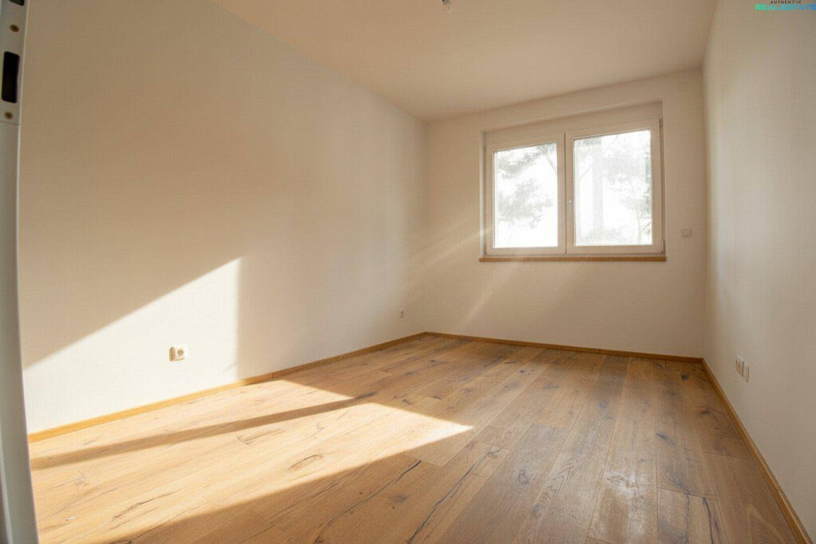Helles, leeres Schlafzimmer mit Holzboden und einem großen Fenster, das viel Sonnenlicht bietet.