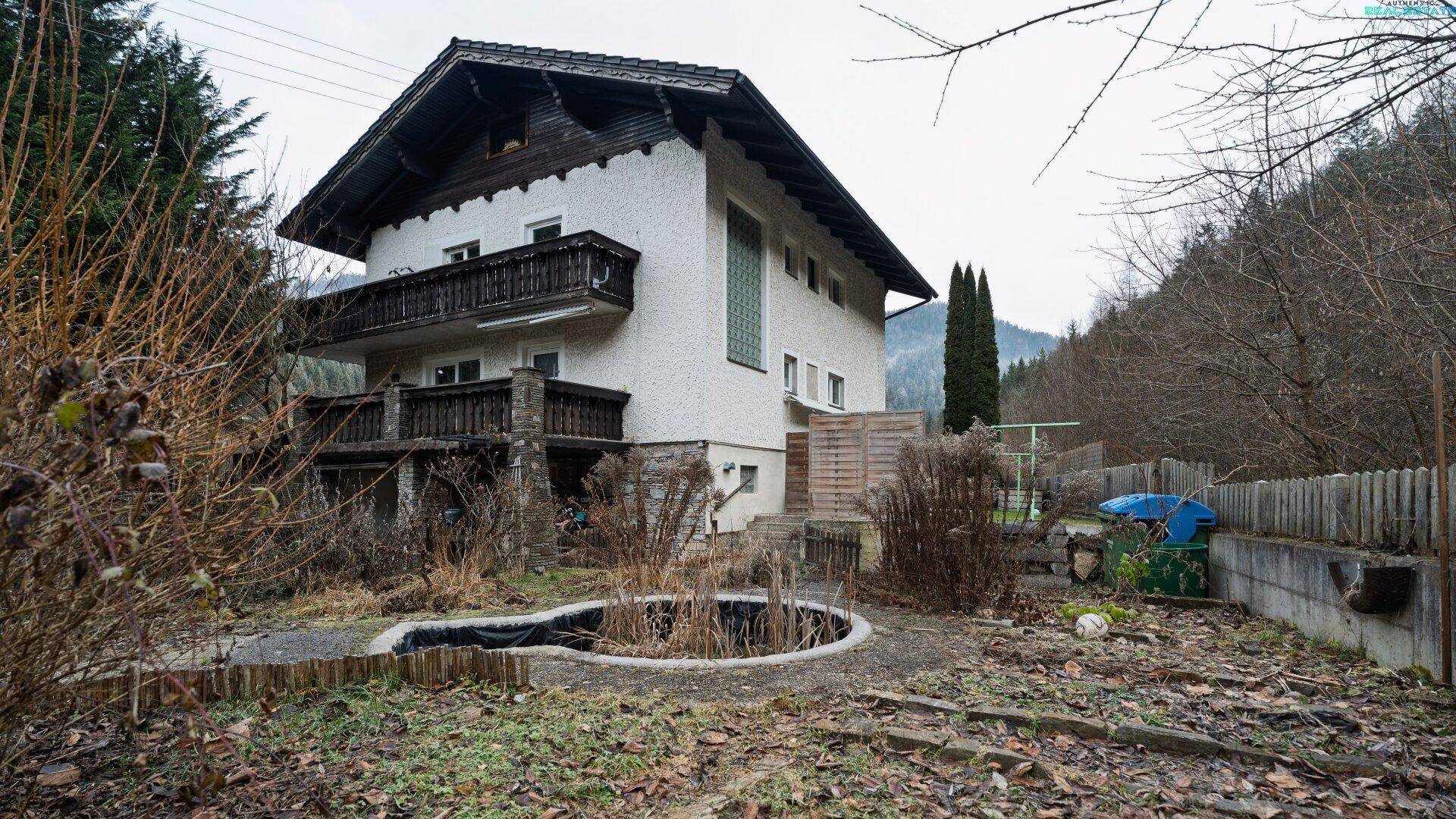 Haus mit Gartenbereich, Teich und Blick auf die bewaldete Umgebung.