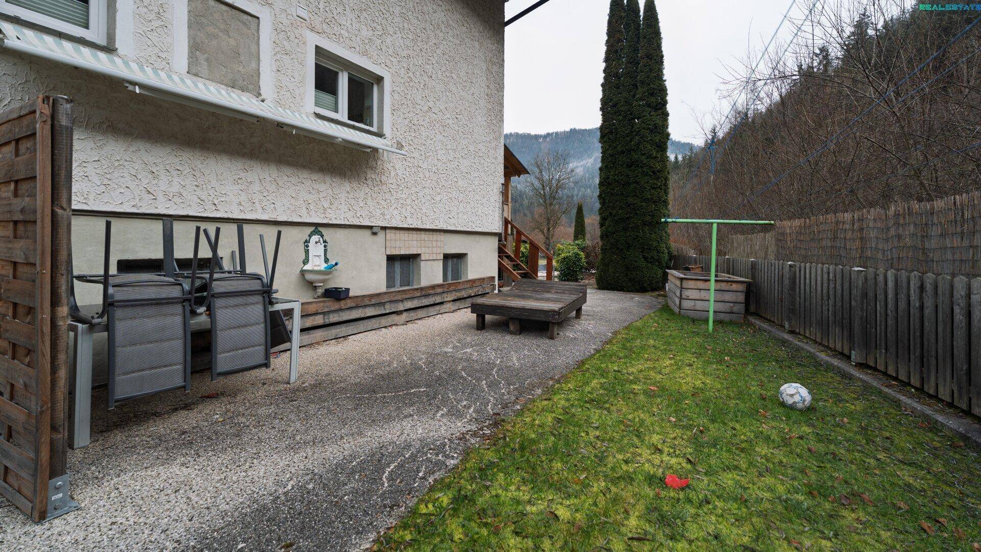 Gartenbereich neben dem Haus mit gepflastertem Weg, Rasenfläche und Blick auf die umliegende Natur.