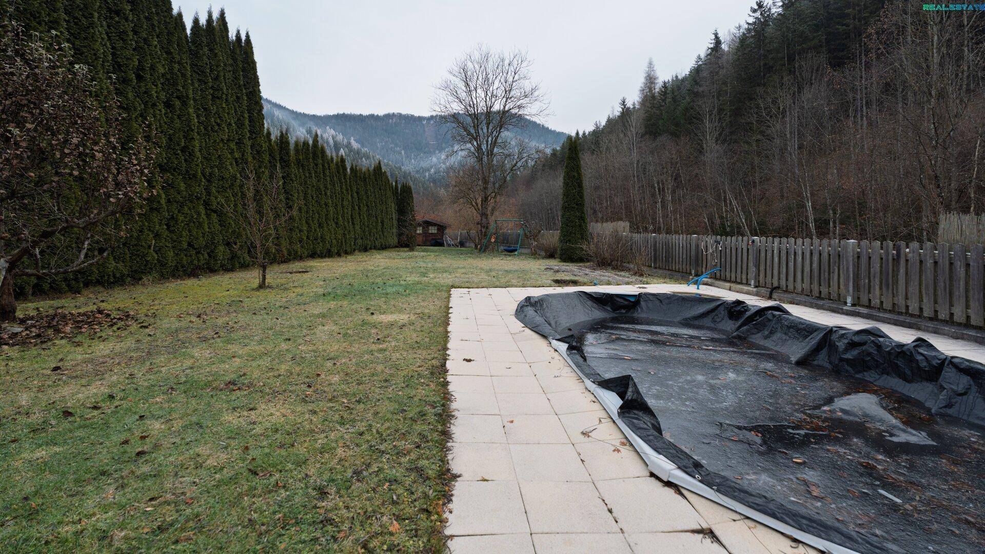 Weitläufiger Garten mit Rasen, umzäuntem Pool und hohen Hecken, umgeben von Natur.