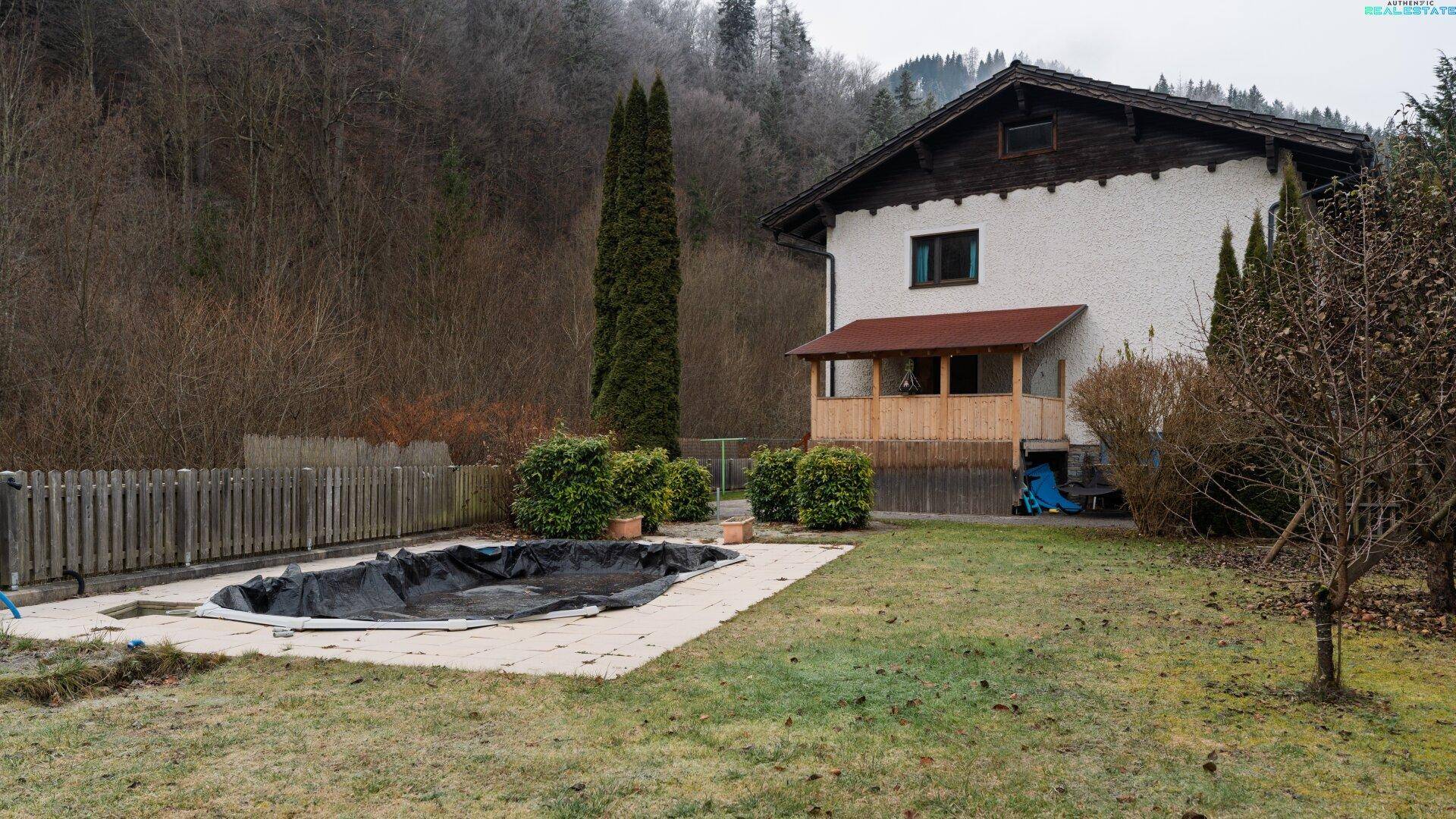 Hausfassade mit überdachtem Balkon und großem Gartenbereich inklusive Pool und Hecken.