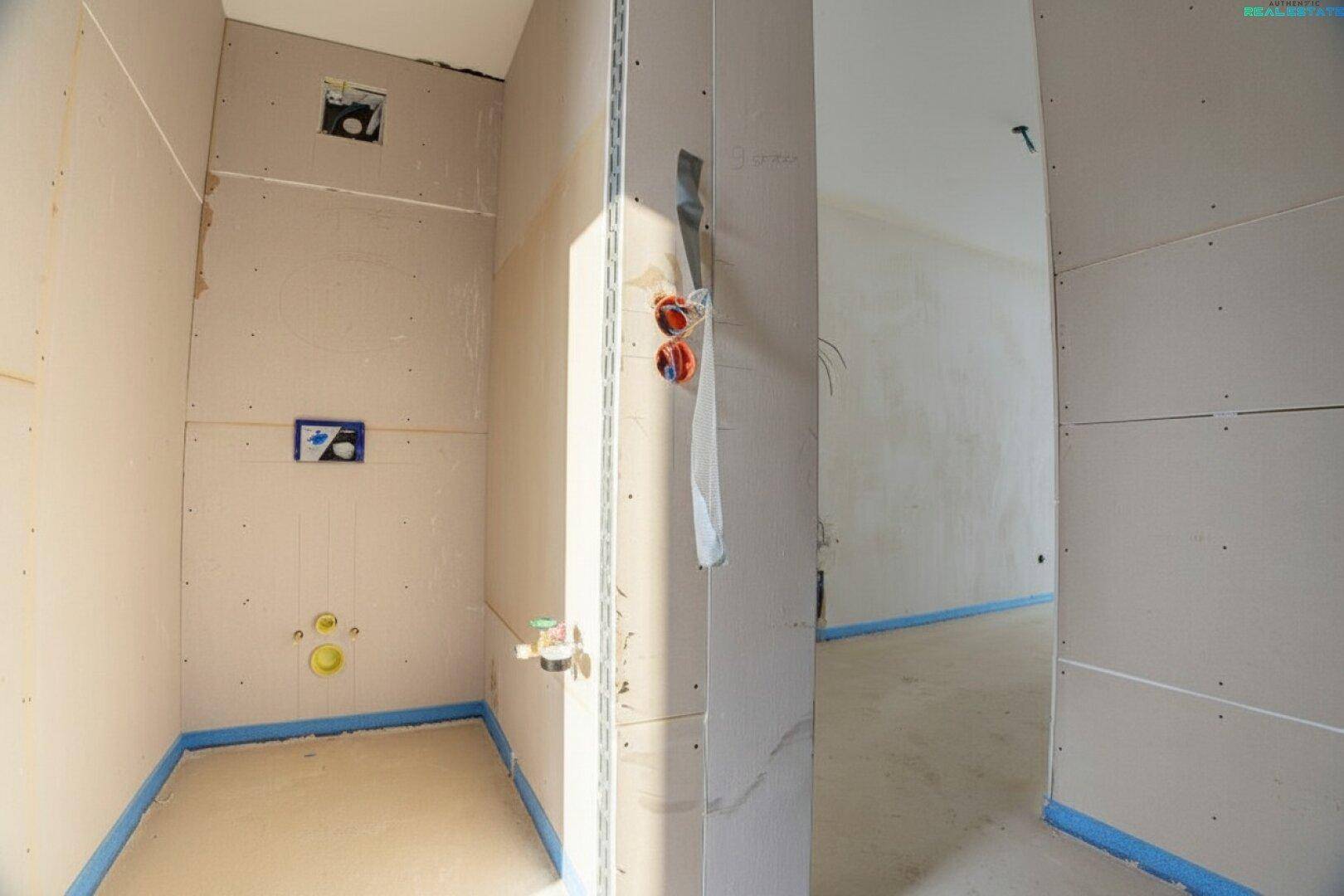 Badezimmer im Rohbau mit Trockenbauwänden und sichtbaren Installationen für Toilette und Waschbecken.
