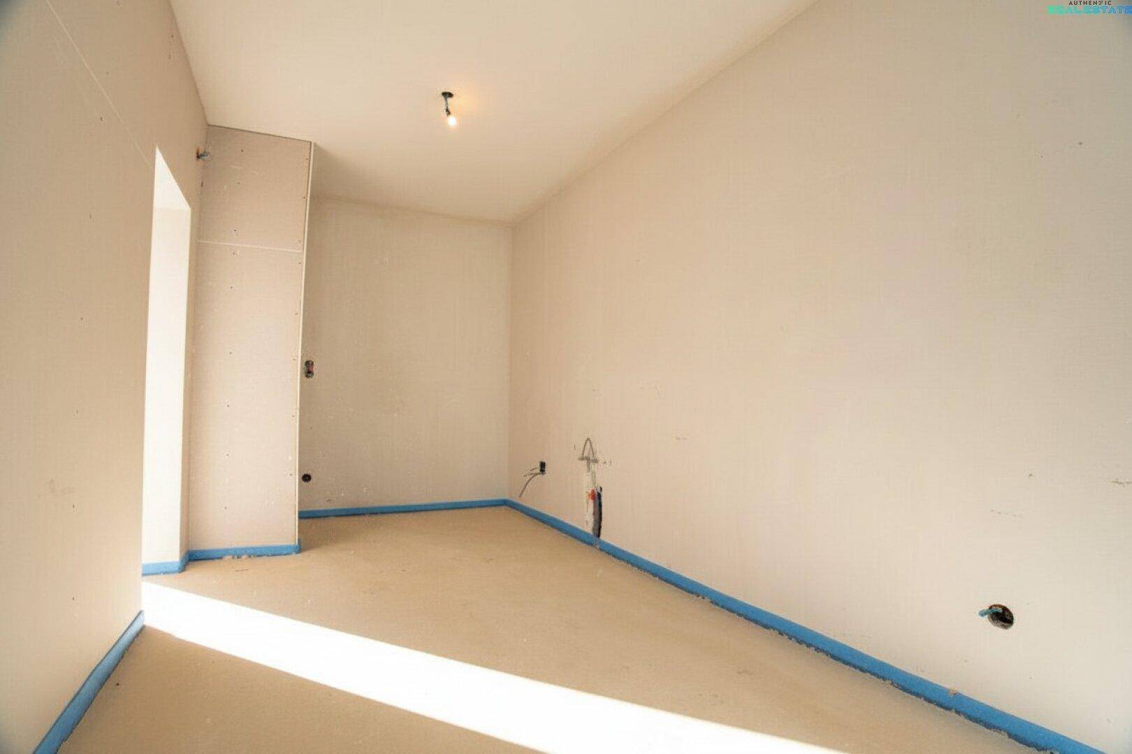 Küchenbereich im Rohbau mit vorbereiteten Anschlüssen und viel Tageslicht durch das Fenster.