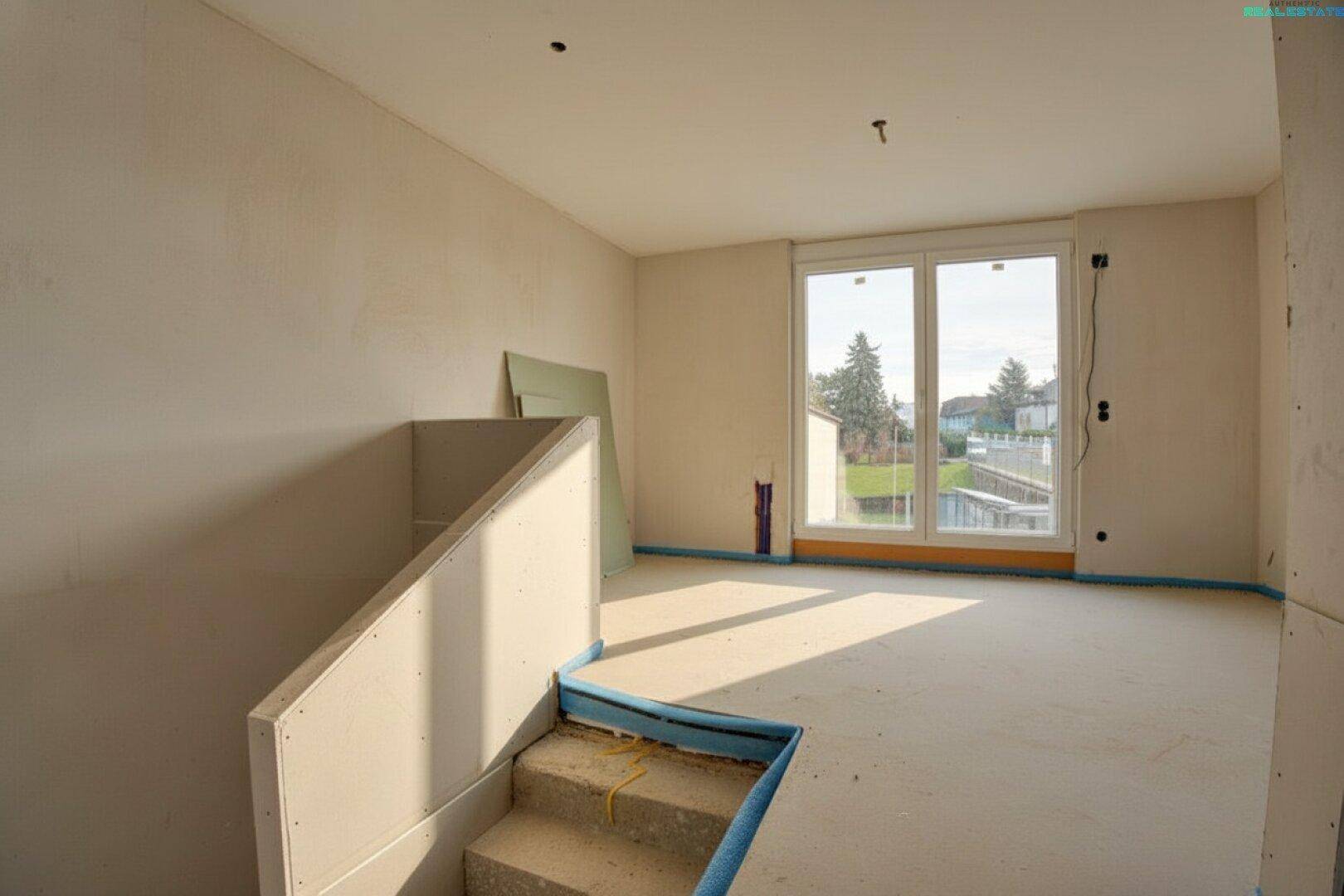 Wohnbereich im Rohbau mit Treppenaufgang und großem Fenster, das viel Tageslicht hereinlässt.