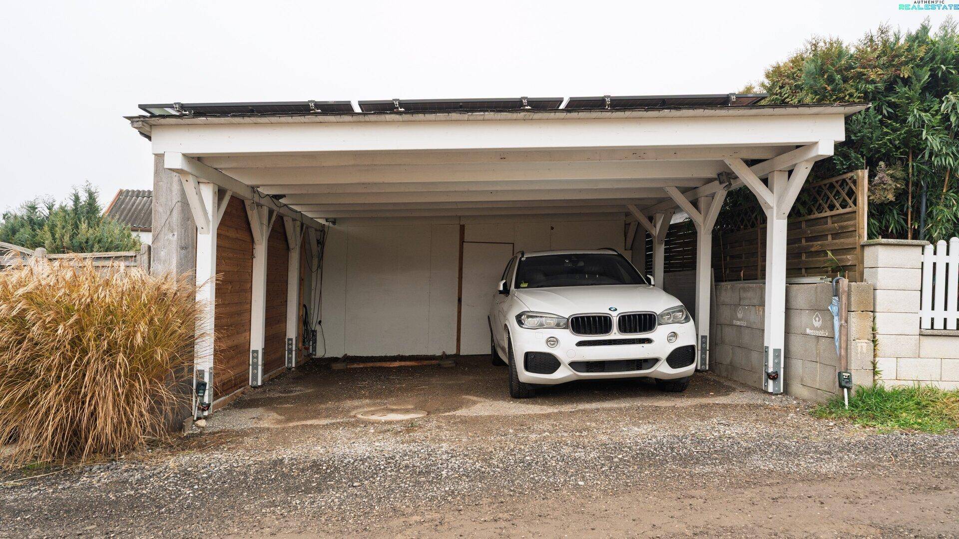 Geräumiger Carport mit Holzbalken, der Platz für ein Fahrzeug bietet und von Grün umgeben ist.