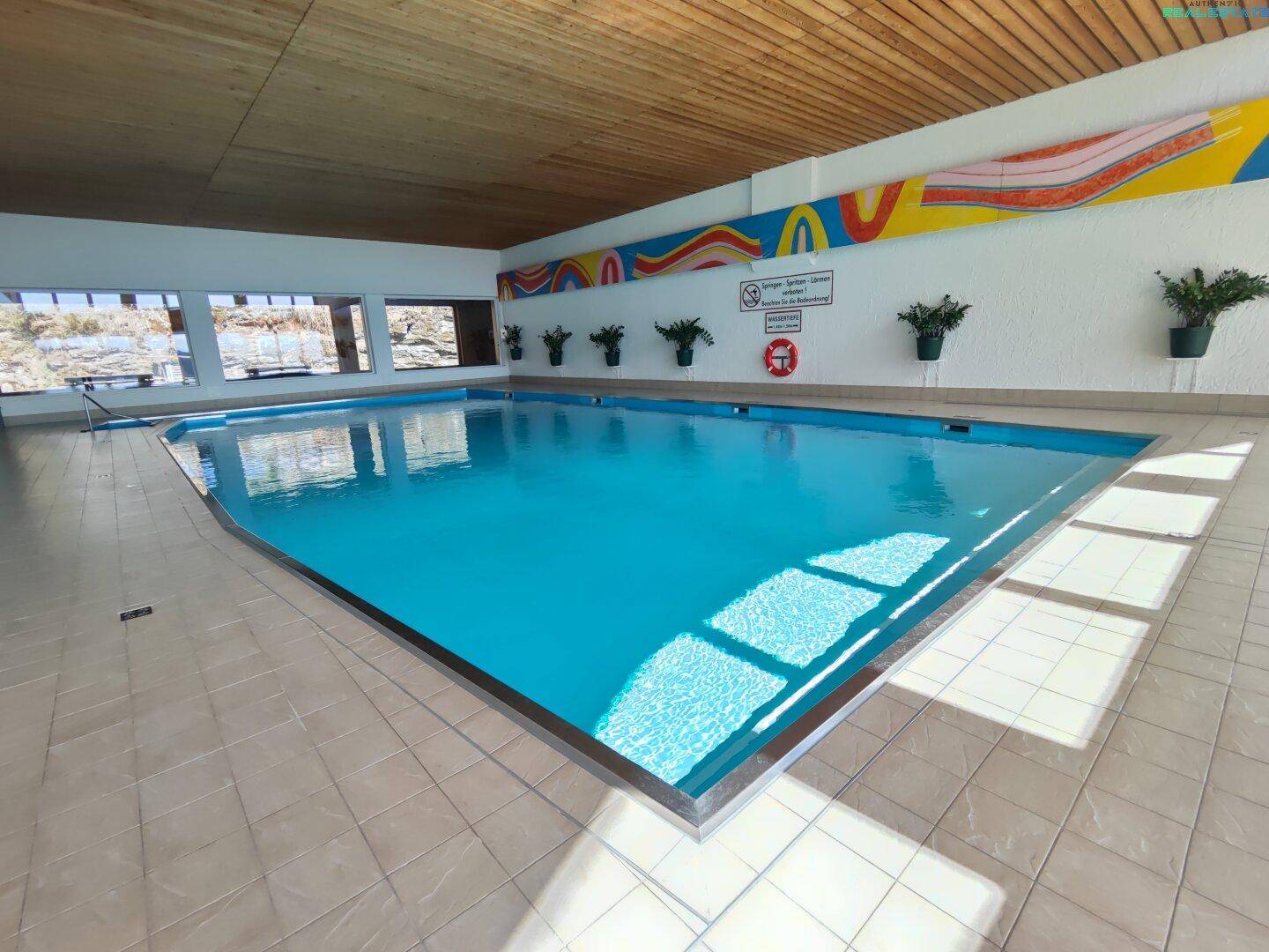 Großes Hallenbad mit hellen Fliesen und großen Fenstern, ideal zum Schwimmen und Entspannen.