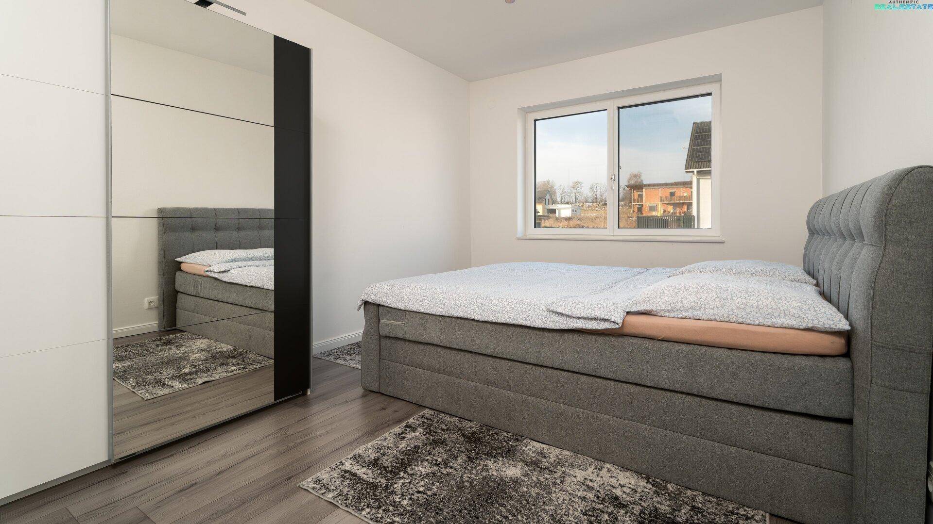 Helles Schlafzimmer mit grauem Boxspringbett, Holzboden und großem Fenster.