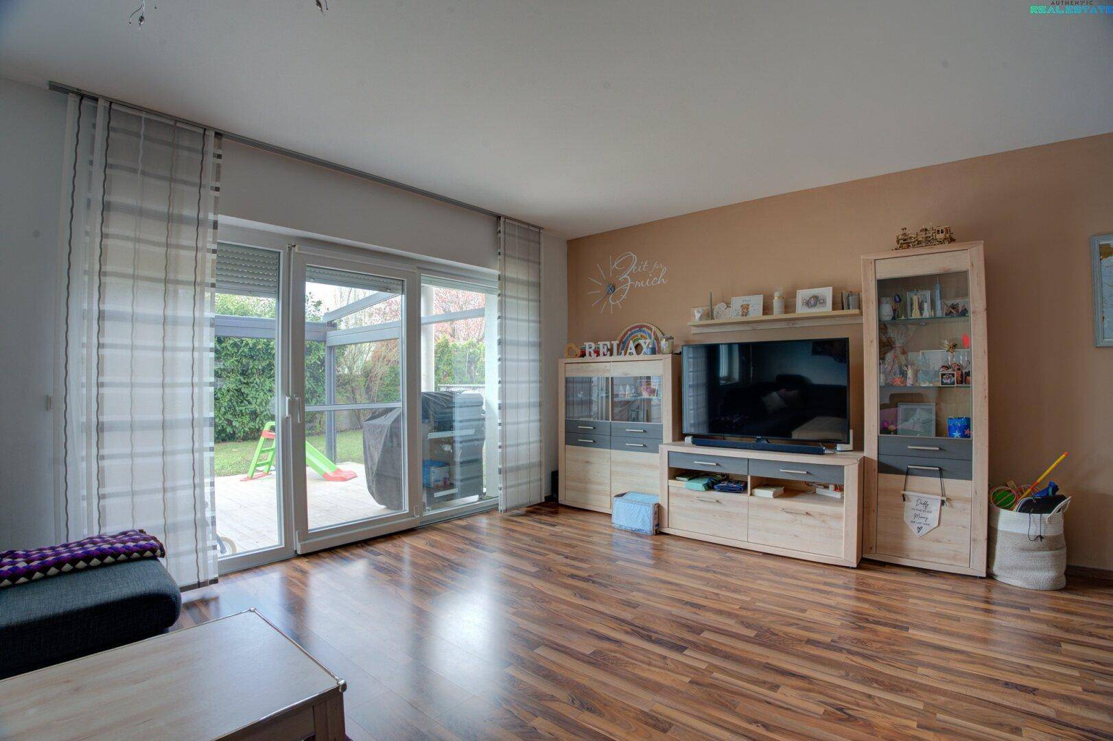 Heller Wohnbereich mit TV-Möbeln, Holzboden und großem Fenster mit Blick auf den Garten.