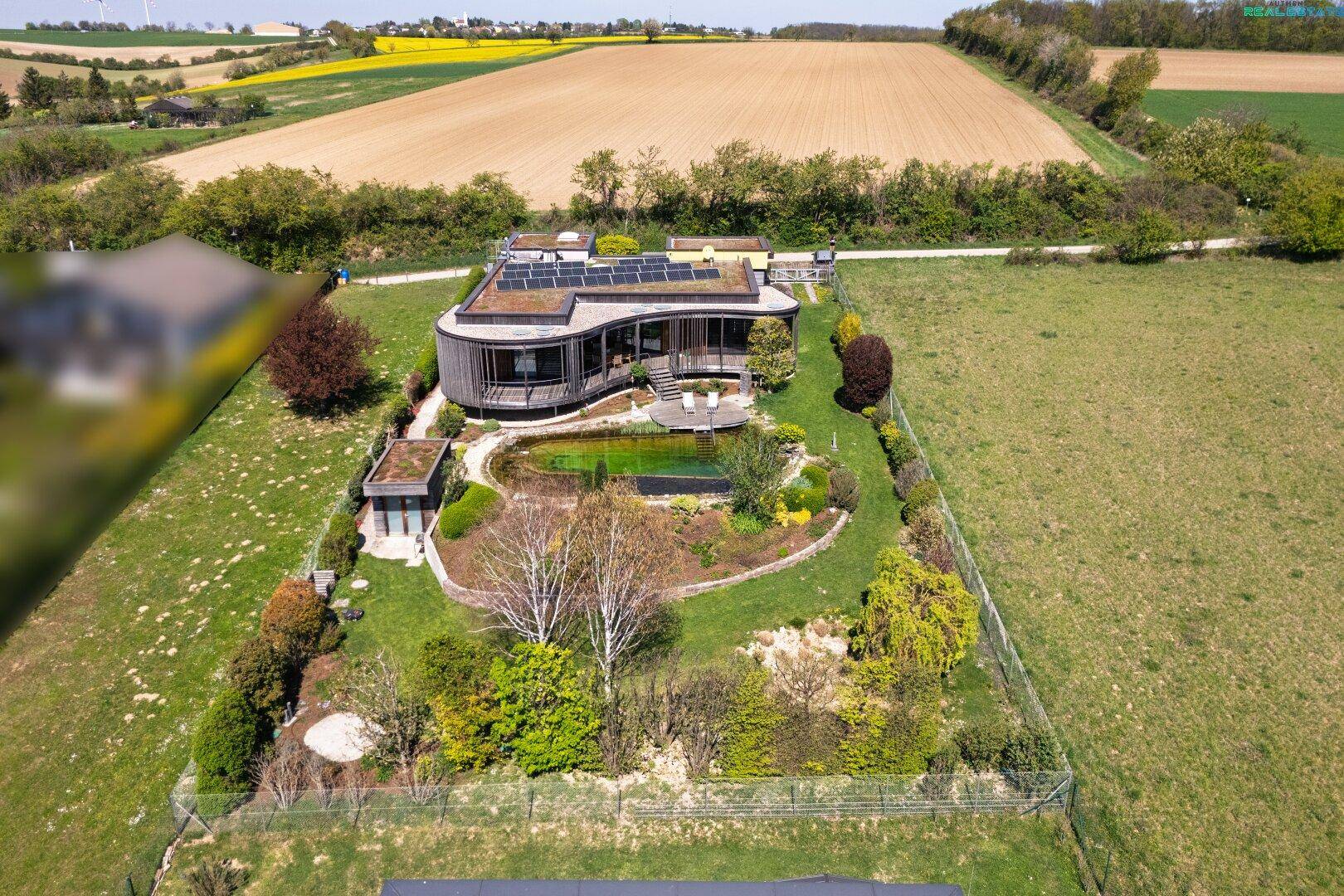 Weitläufige Ansicht des Anwesens mit modernem Haus, Naturpool und angrenzenden Feldern unter blauem Himmel.
