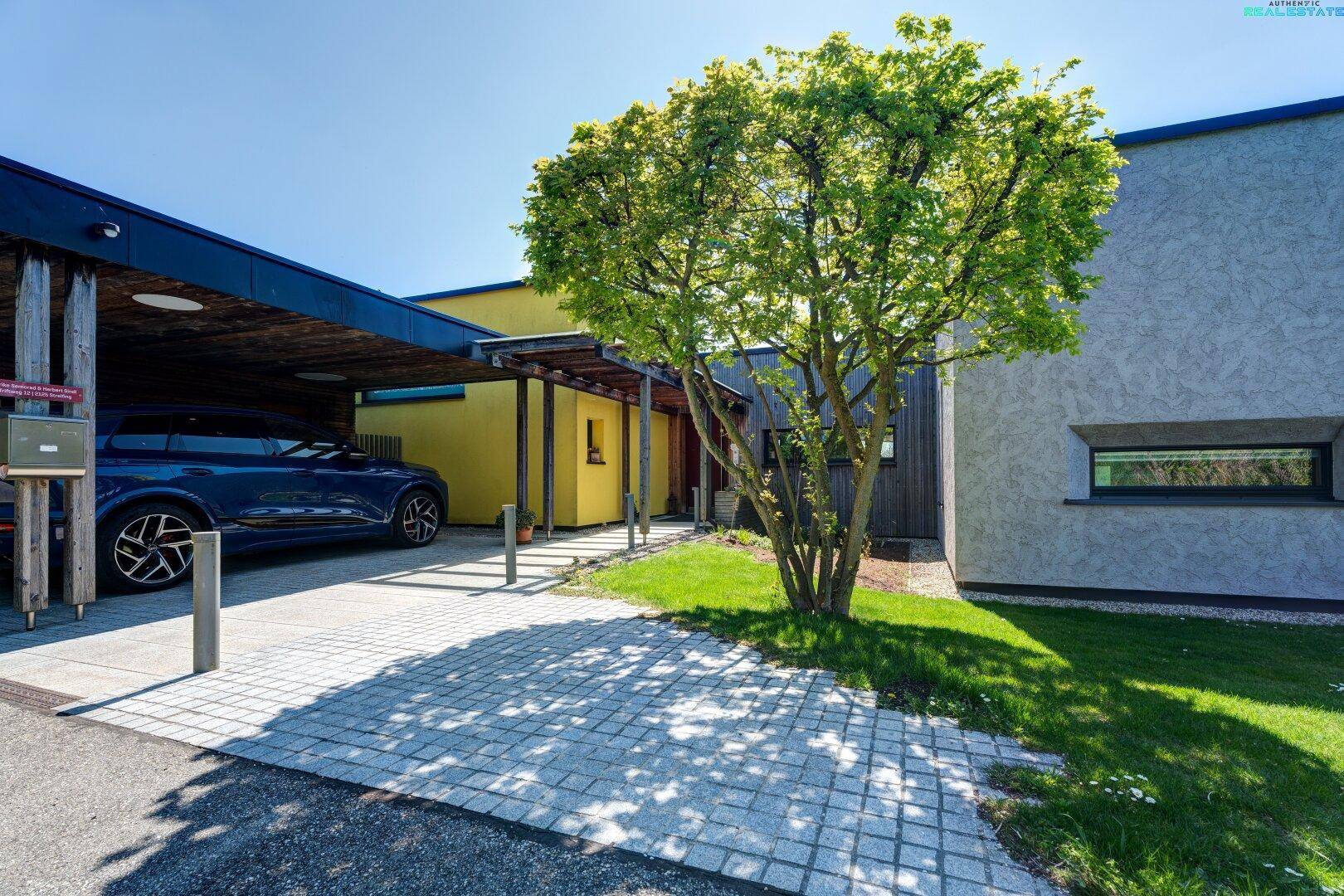 Eingangsbereich des Hauses mit Carport und gepflastertem Weg unter einem Baum.