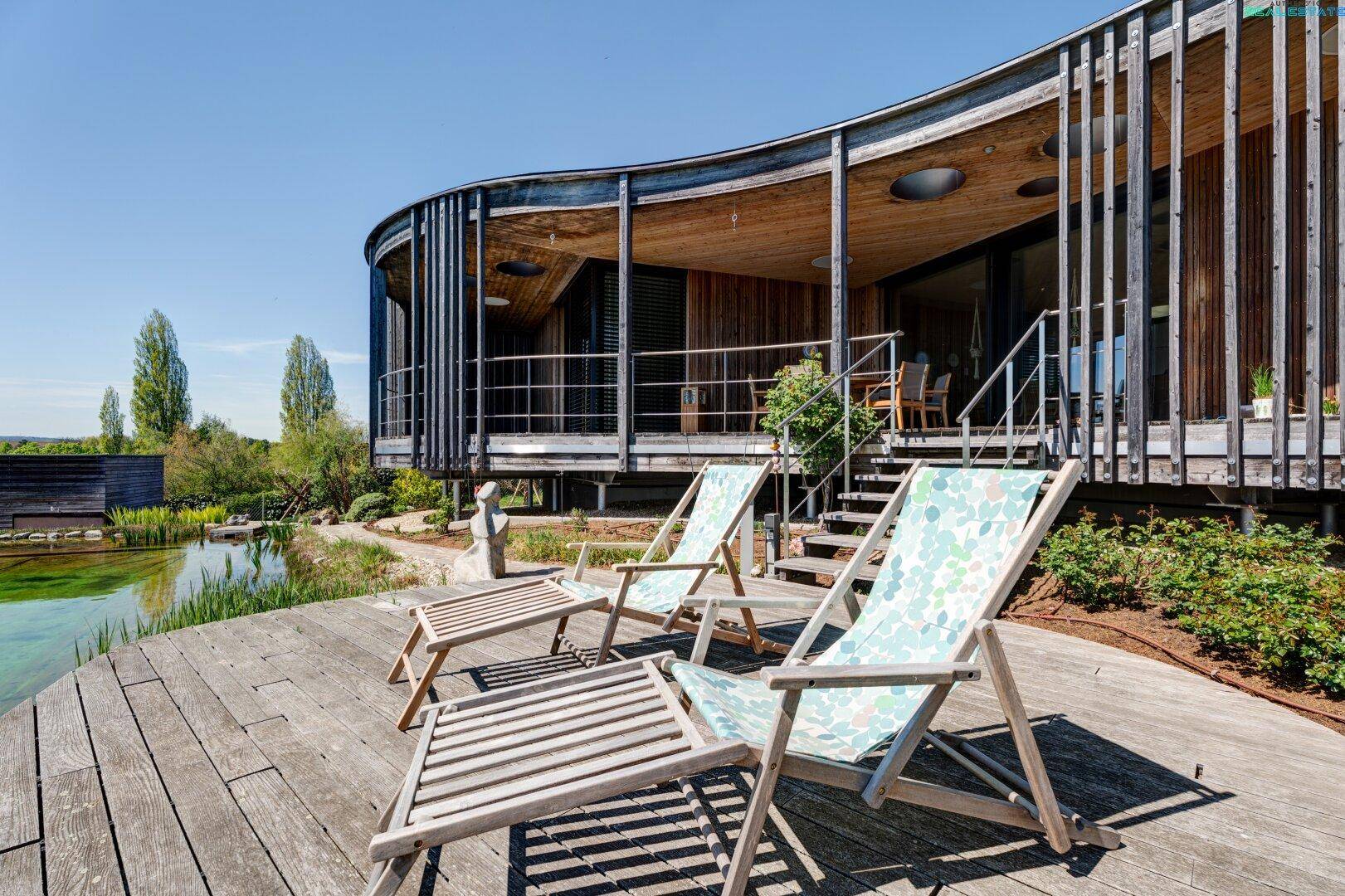 Großzügiges Holzdeck mit Liegestühlen und Blick auf den Naturpool und die moderne Holzfassade.