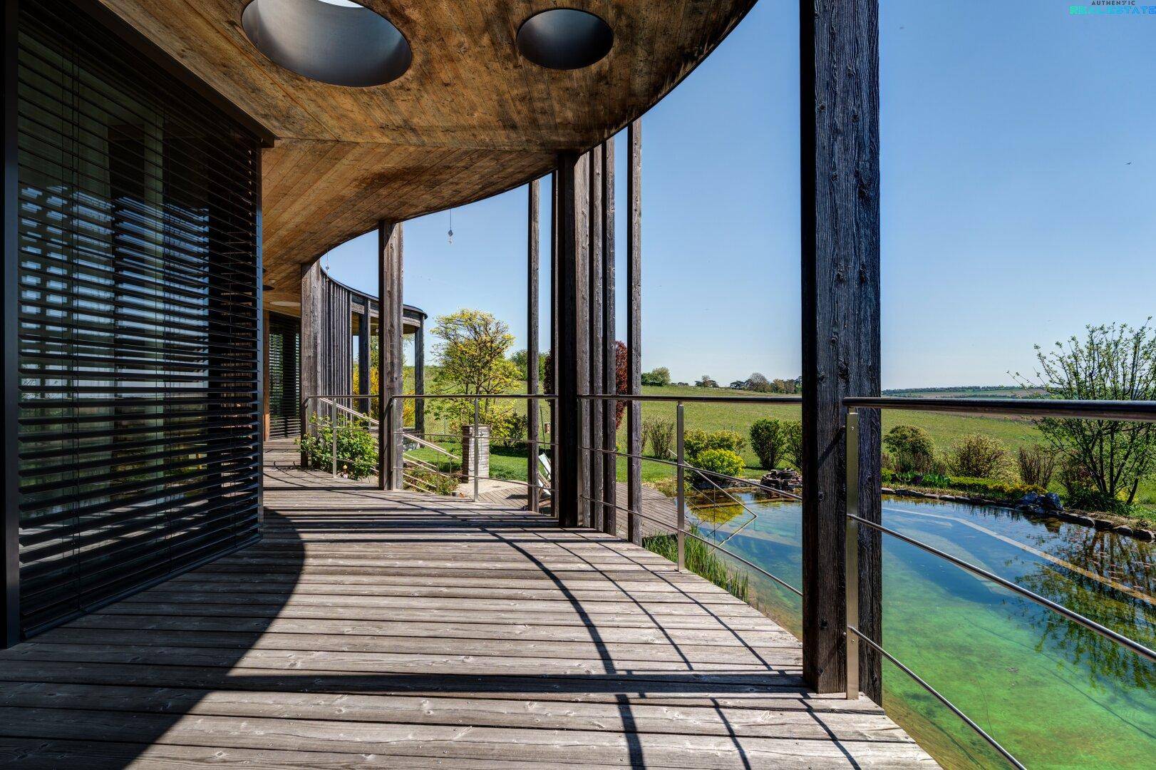 Überdachte Holzterrasse mit weitem Blick über den Naturpool und die grüne Landschaft.