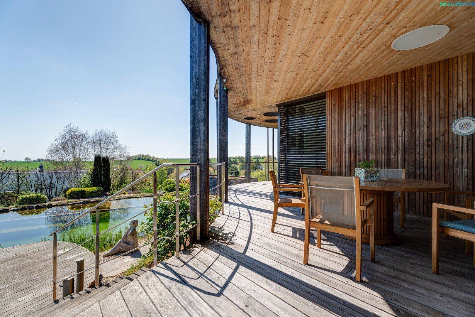 Überdachte Holzterrasse mit Blick auf den Naturpool und die umliegende grüne Landschaft.
