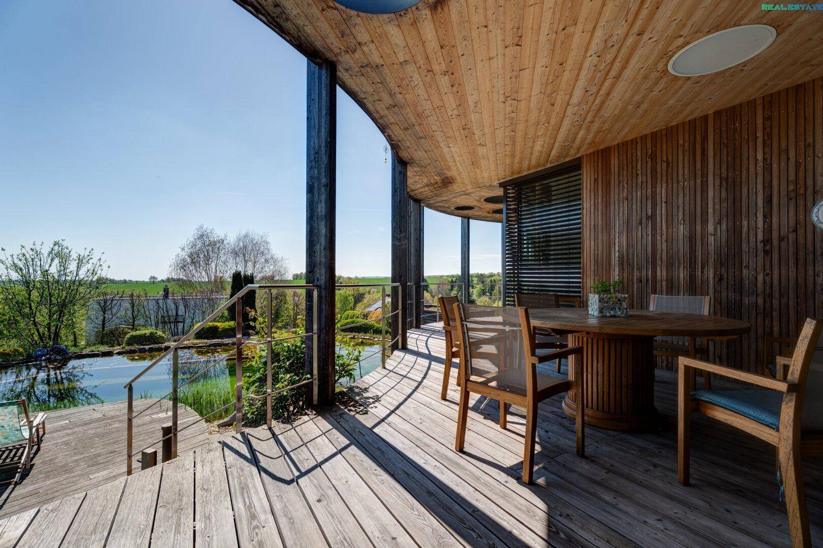 Geräumige Holzterrasse mit Essbereich und weitem Blick über den Naturpool und die Landschaft.
