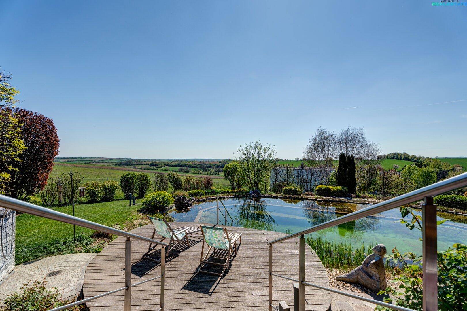 Holzdeck mit Liegestühlen am Naturpool und weitem Blick über die grüne Landschaft.