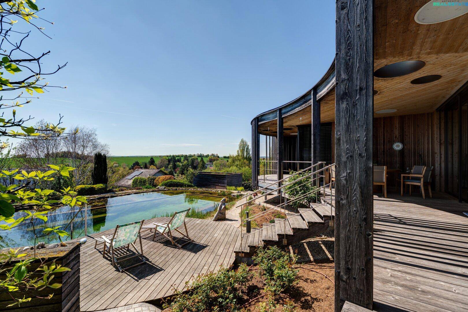 Großzügiges Holzdeck mit Liegestühlen am Naturpool und der geschwungenen Holzfassade des Hauses.