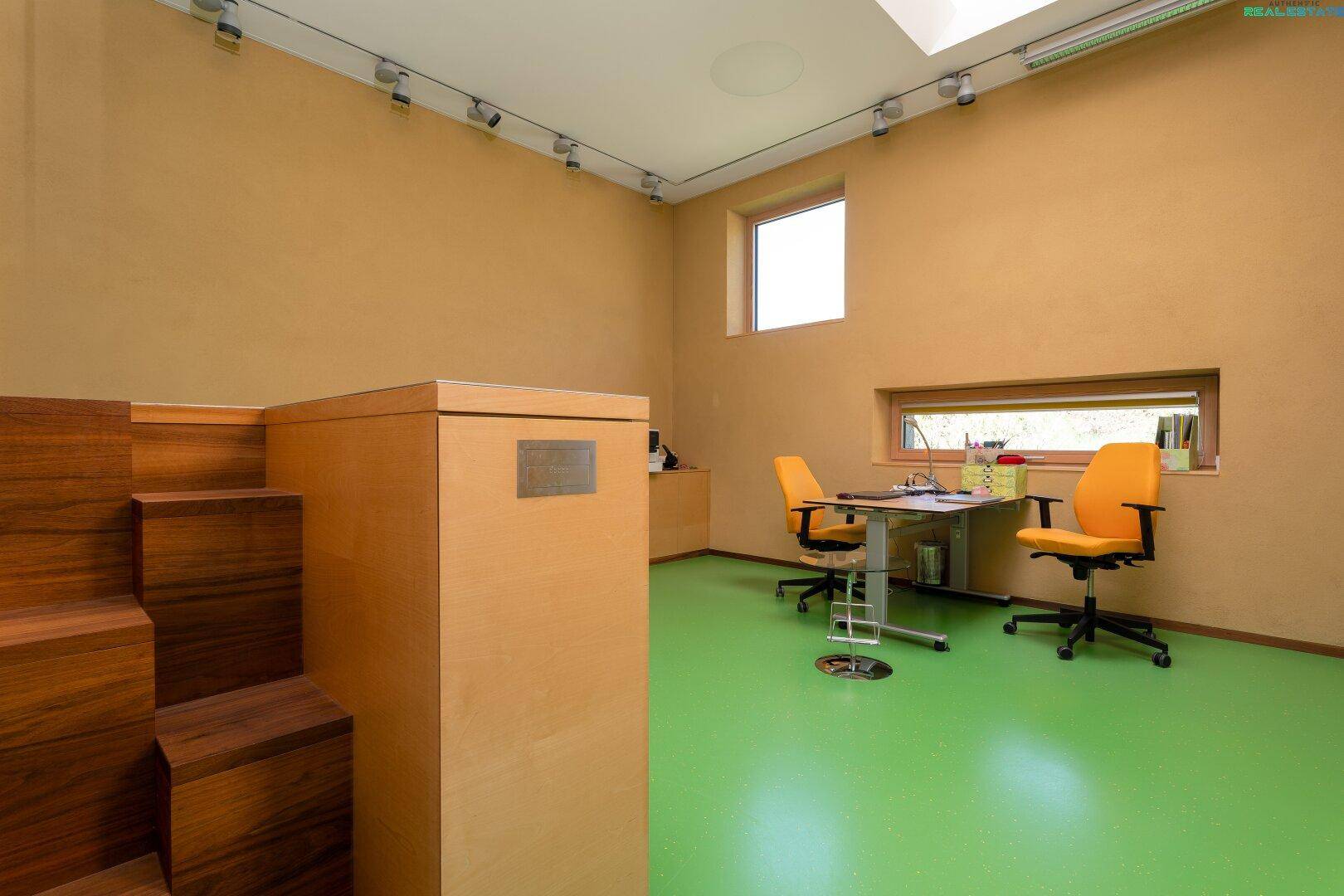 Helles Büro mit grünem Boden, Schreibtisch und Blick durch die Fenster ins Grüne.