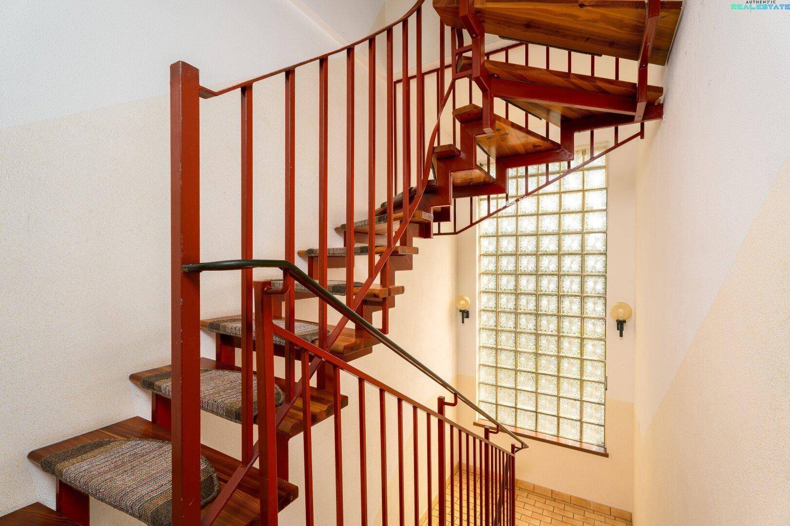 Wendeltreppe mit Holzstufen und rotem Geländer, beleuchtet durch ein großes Glasbausteinfenster.