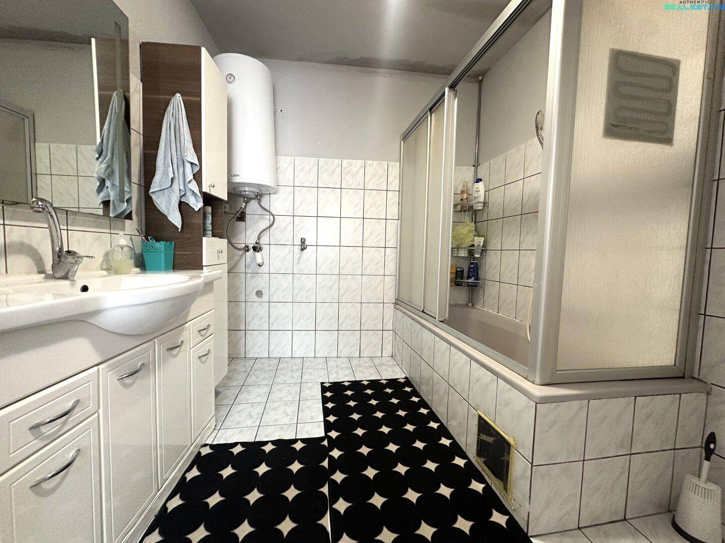 Badezimmer mit Fliesenboden, Badewanne mit Duschabtrennung und Einzelwaschbecken.