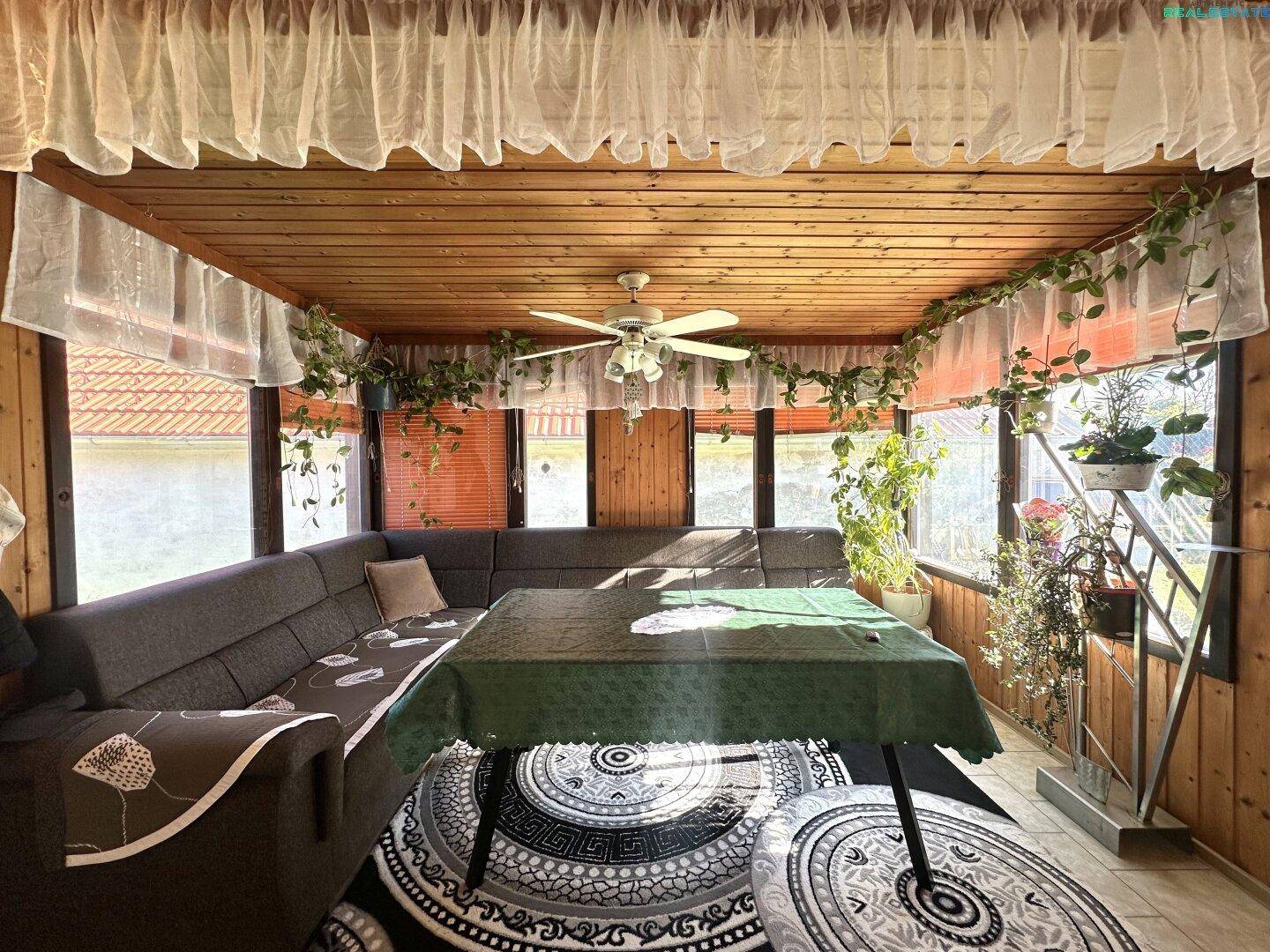 Gemütlicher Wintergarten mit großen Fenstern, Holzverkleidung und Deckenventilator.