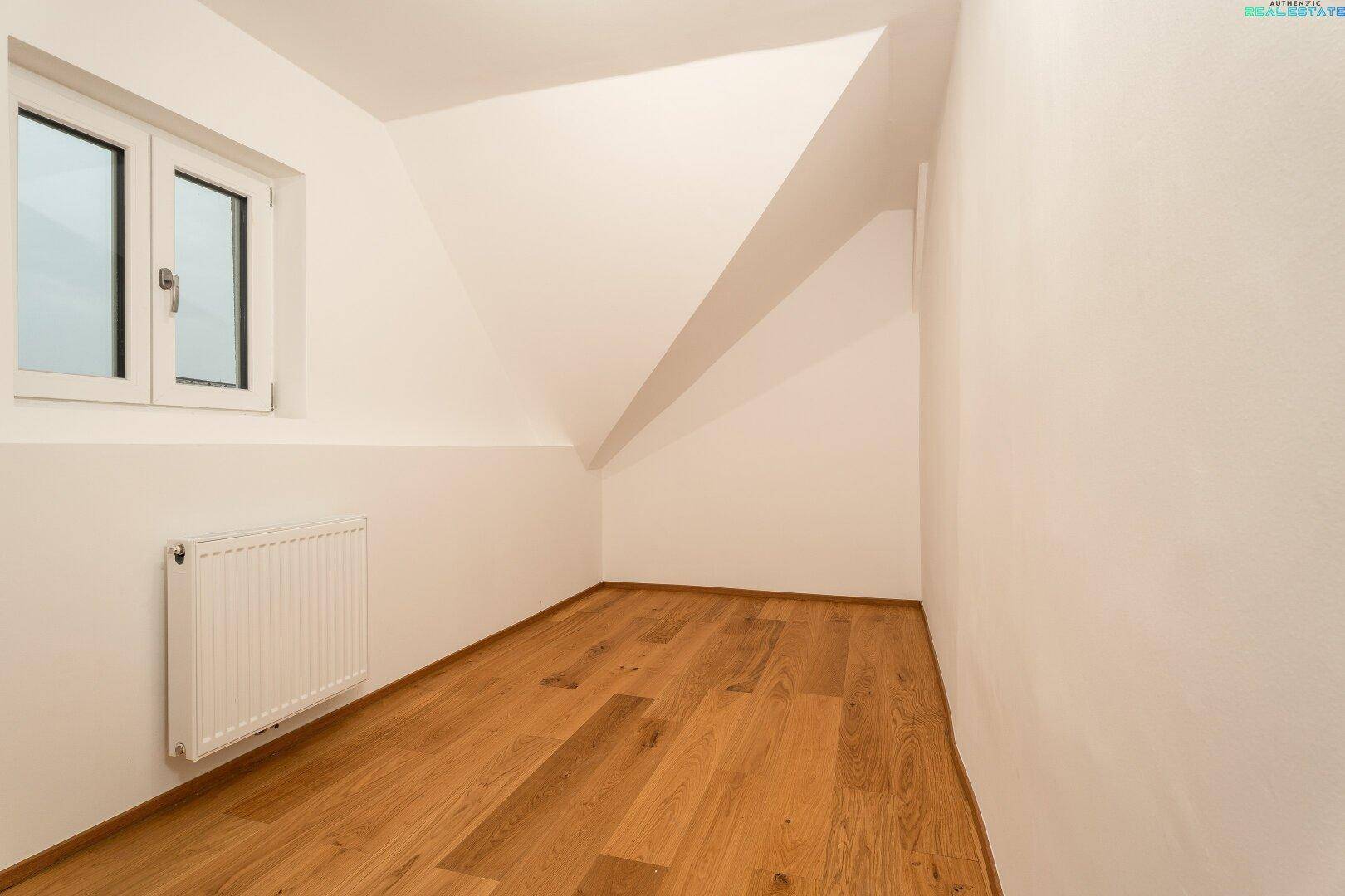 Kleines Dachzimmer mit schrägen Wänden, Holzfußboden und Fenster.
