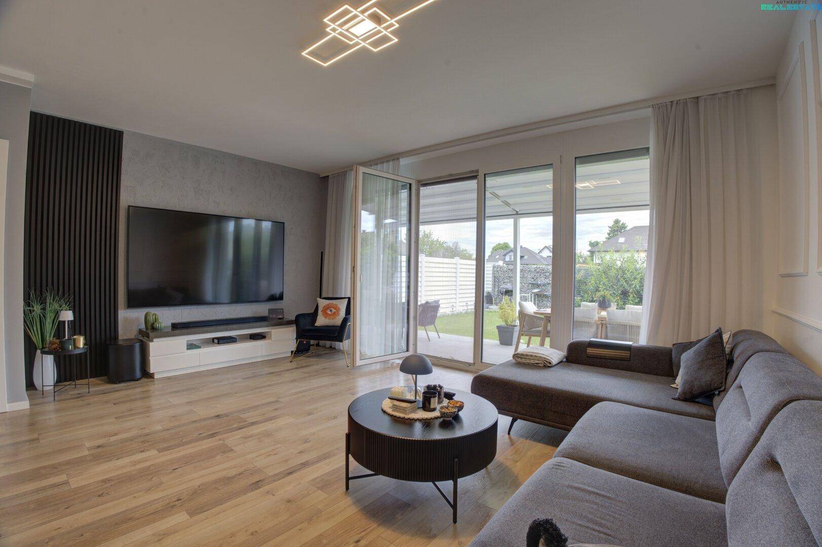 Modern eingerichtetes Wohnzimmer mit bequemer Couch, Holzboden und viel Tageslicht durch große Fenster.