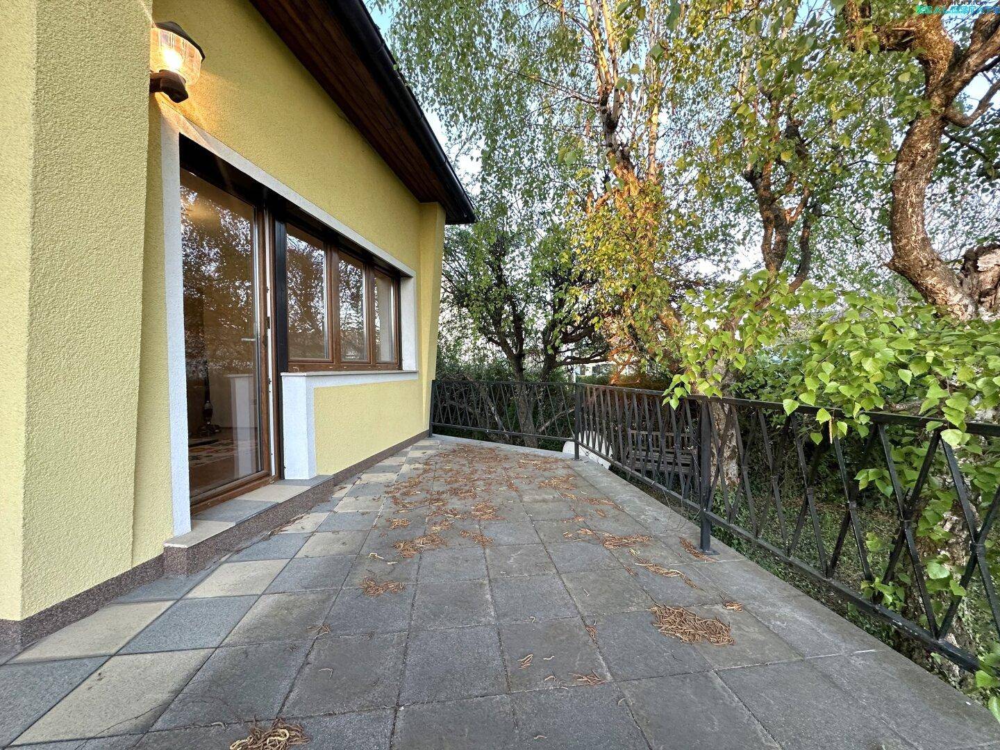 Lange, geflieste Terrasse mit Metallgeländer und umgebenden Bäumen.