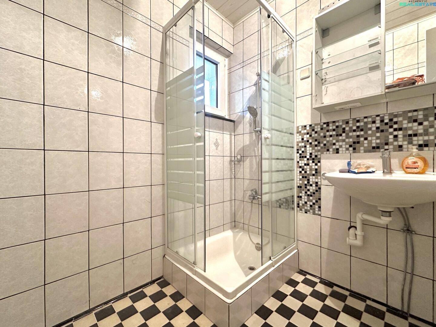 Badezimmer mit Duschkabine, Waschbecken und Fenster, hell gefliest.