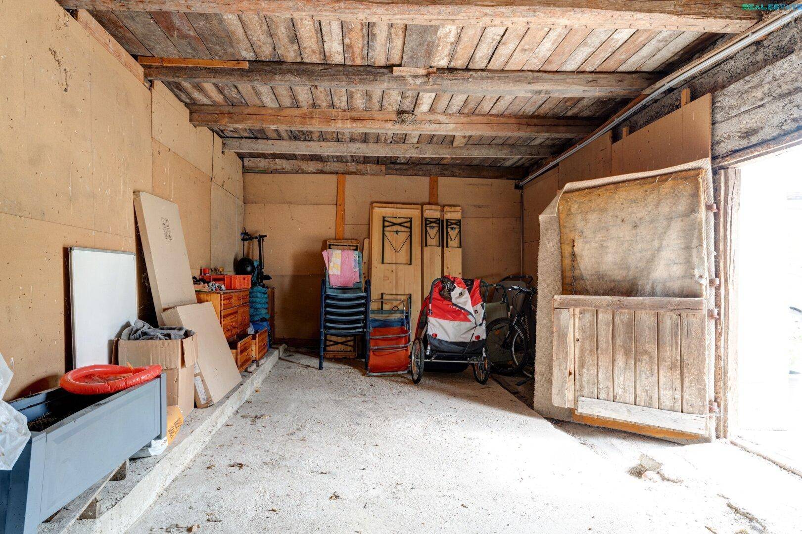 Geräumige Garage oder Werkstatt mit Holztor, Betonboden und sichtbaren Deckenbalken.