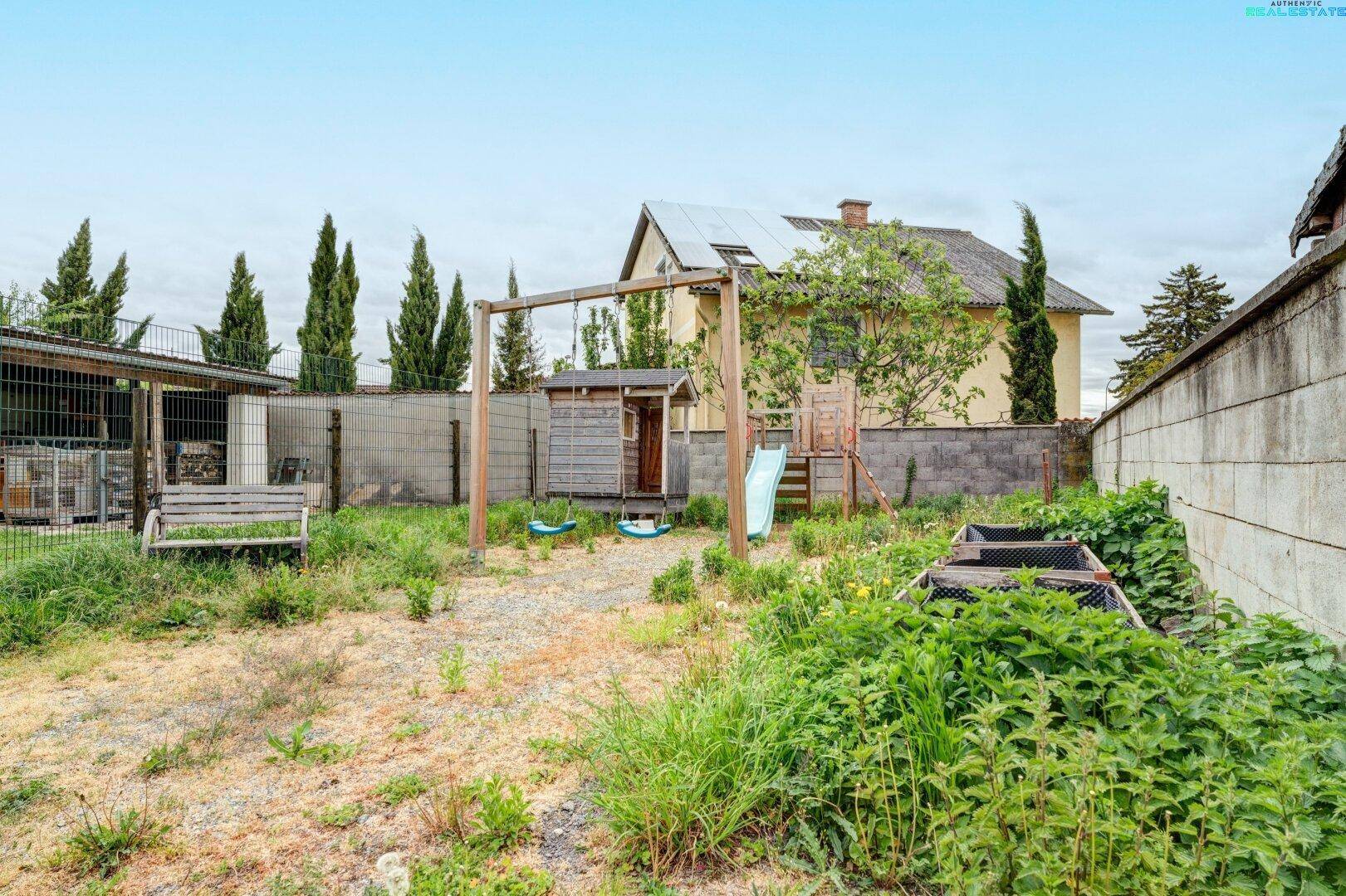 Gartenbereich mit Schaukel, Rutsche und kleinem Spielhaus, umgeben von viel Grün.