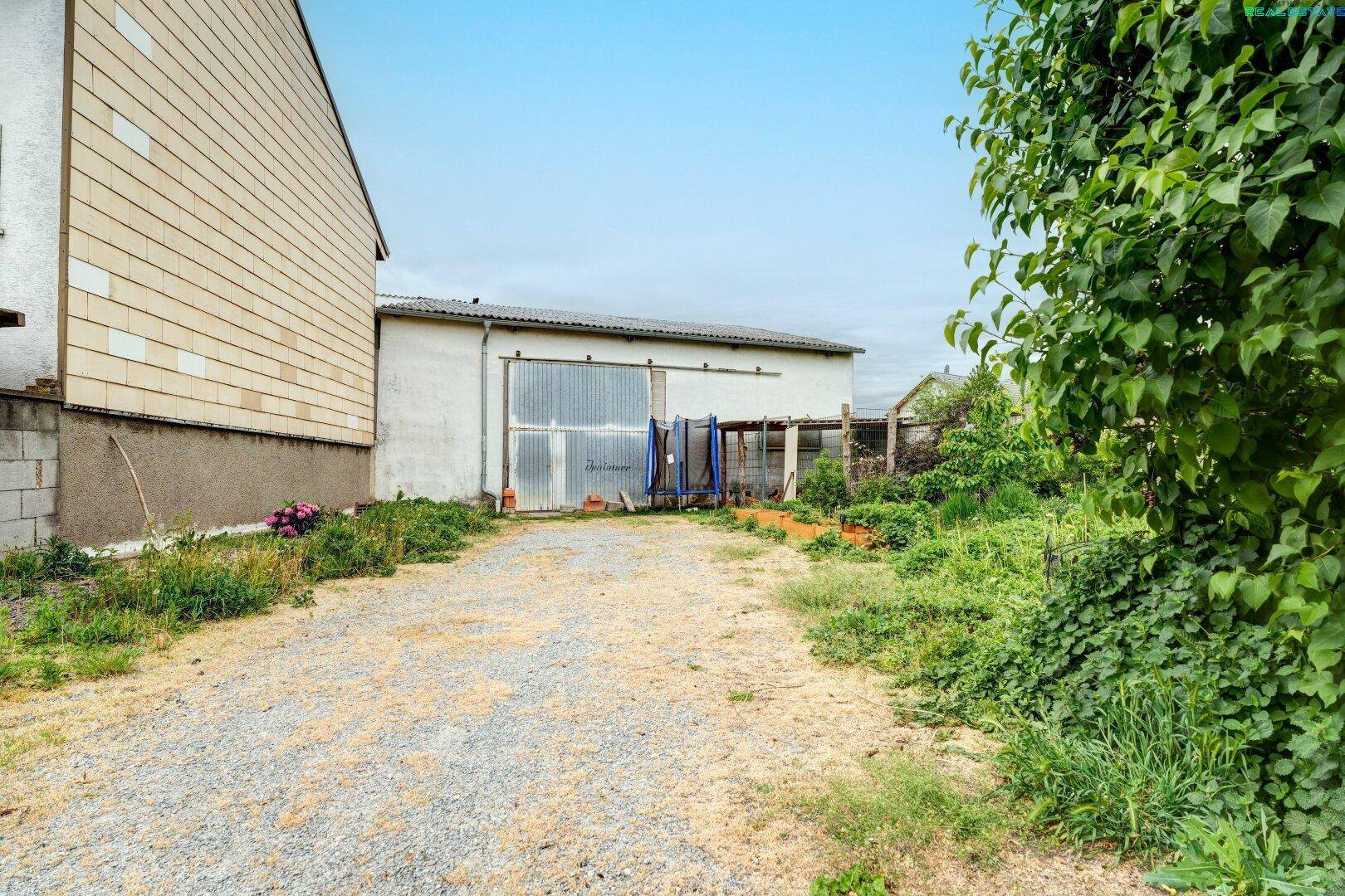 Außenansicht mit Schotterweg, der zu einem großen Garagengebäude führt, umgeben von Vegetation.