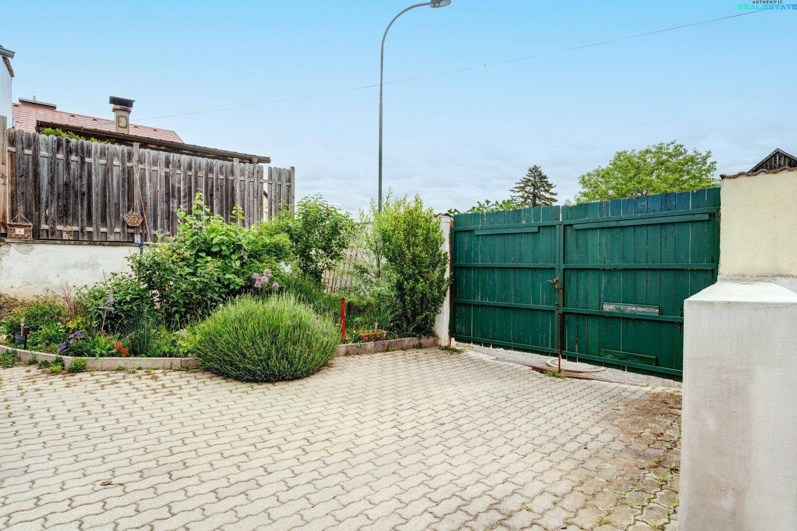 Eingangsbereich mit Pflastersteinen, einem großen grünen Holztor und Gartenbüschen.