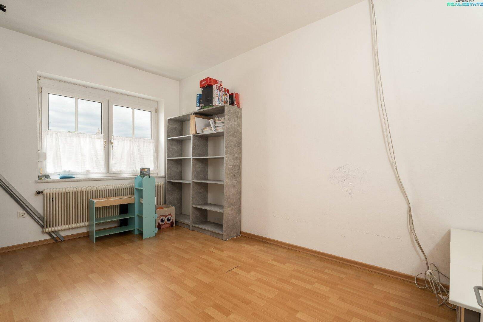Geräumiges Zimmer mit Holzfußboden, großem Fenster, Heizkörper und einem Bücherregal.