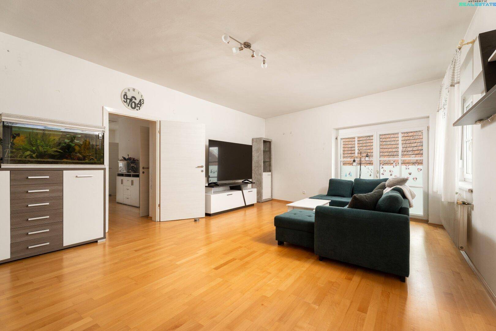 Helles Wohnzimmer mit Holzfußboden, Fernseher, Aquarium und Zugang zu weiteren Räumen.