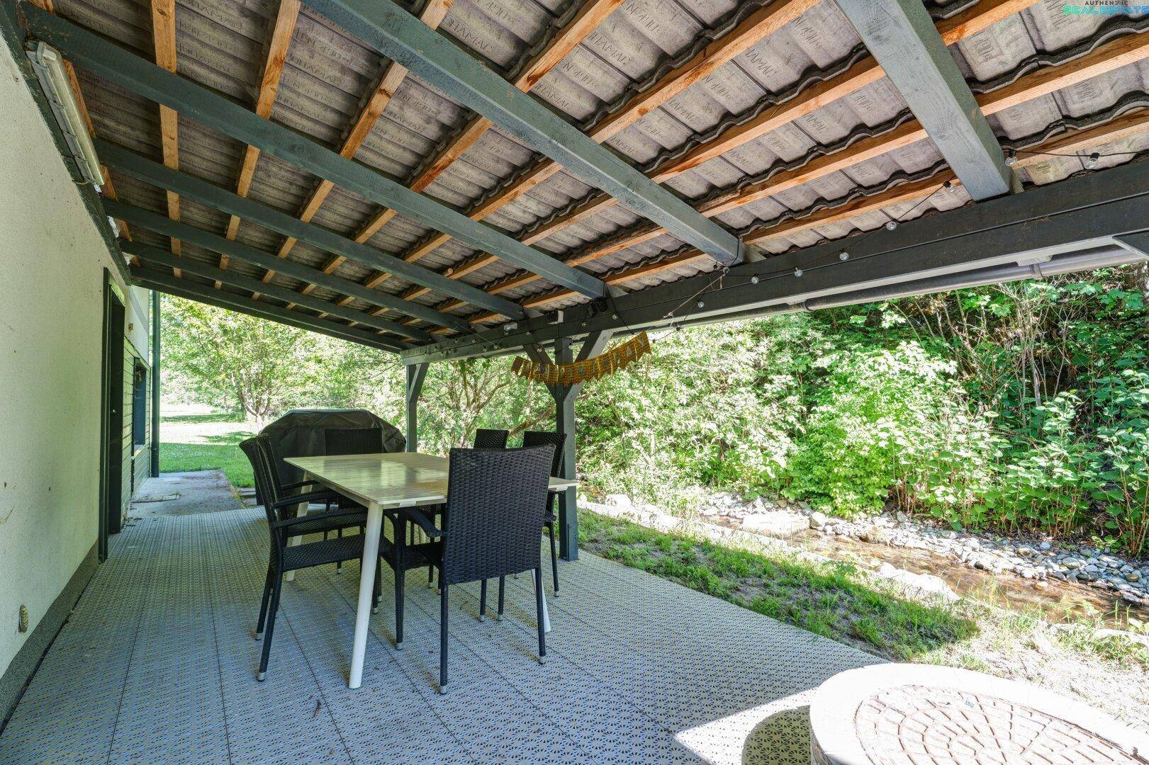 Überdachte Terrasse mit Esstisch und Stühlen, direkt am Bach gelegen.