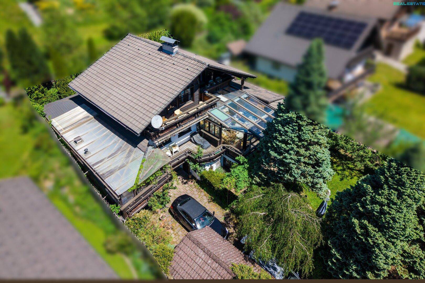Luftaufnahme des Hauses mit Wintergarten, Garten, Parkplatz und umgebender Natur.