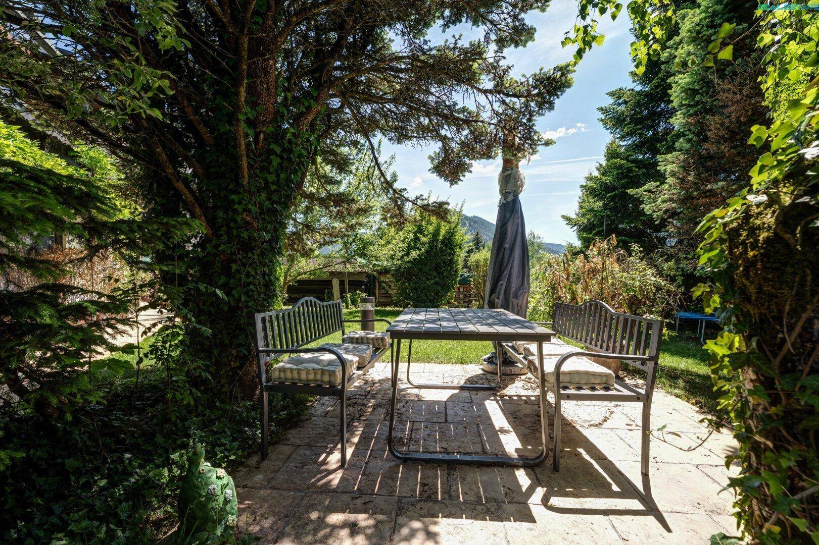 Gemütlicher Außenbereich mit gepflasterter Terrasse, Tisch und Stühlen unter schattenspendenden Bäumen.
