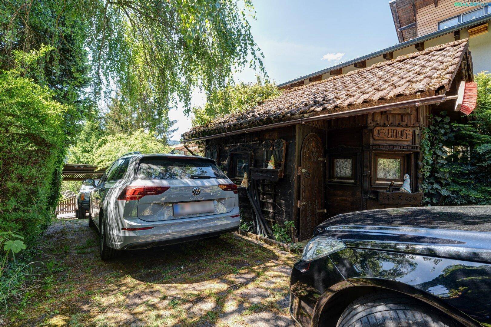 Parkplatz vor einem rustikalen Holzschuppen, umgeben von grüner Hecke und Bäumen.